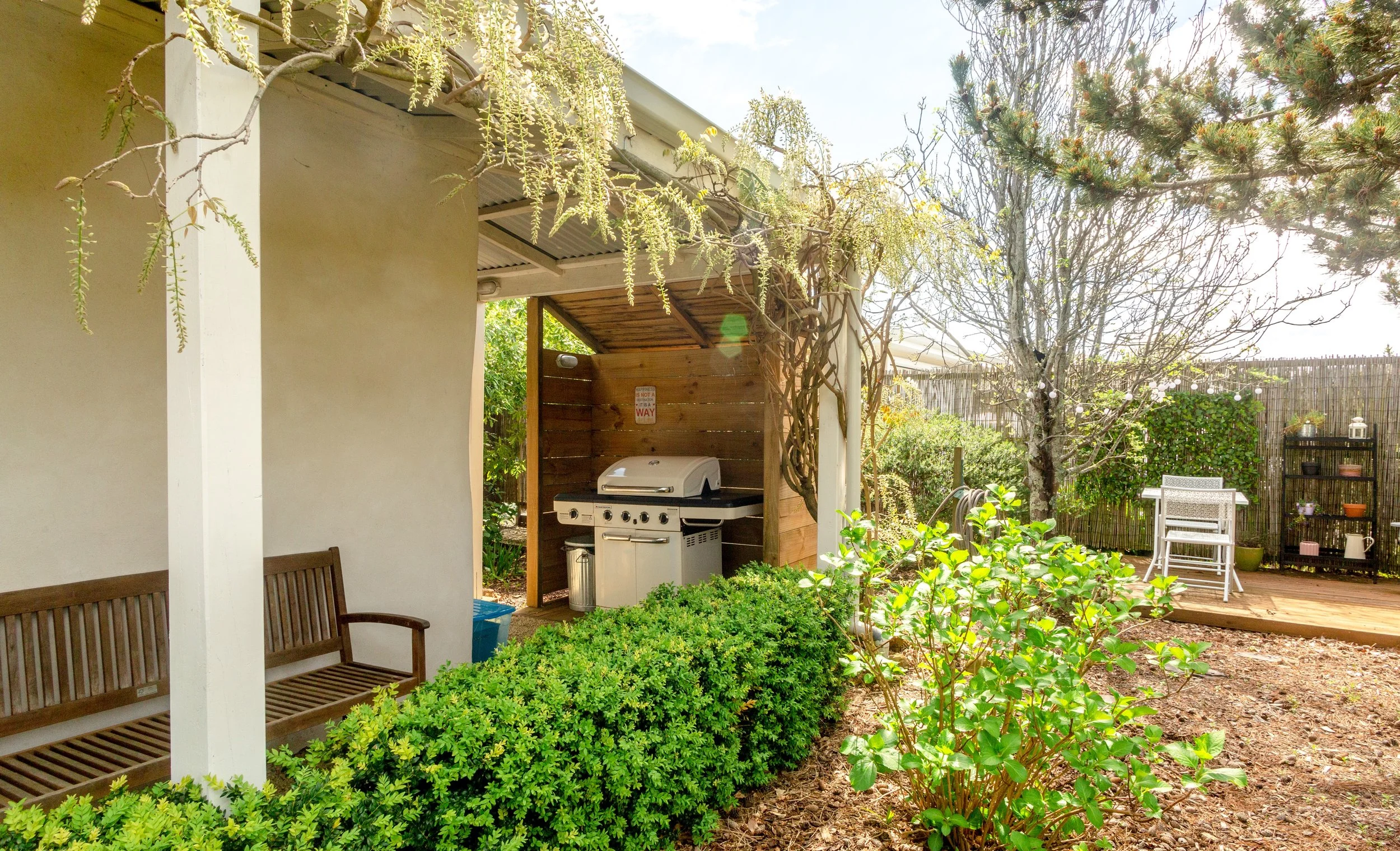 A backyard garden patio with a barbecue grill under a wooden shelter, surrounded by lush green bushes, trees, and outdoor furniture, including a white chair and a small black shelf with potted plants.