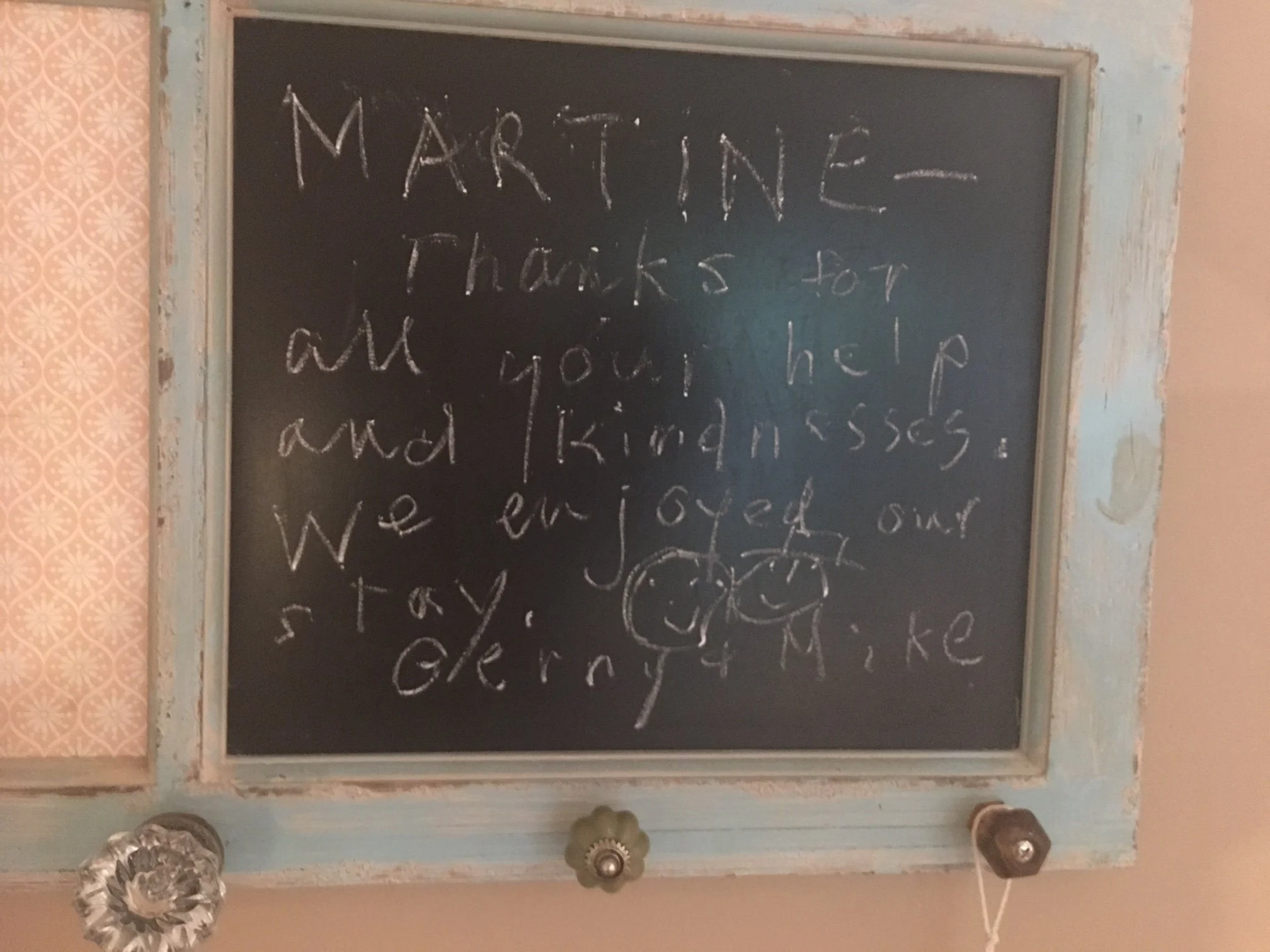 A blackboard with white chalk writing that reads 'MARTINE, Thanks for all your help and kindness!'. The blackboard has a rustic, distressed wood frame. To the left of the blackboard is wallpaper with a pink and white floral pattern. At the bottom of 