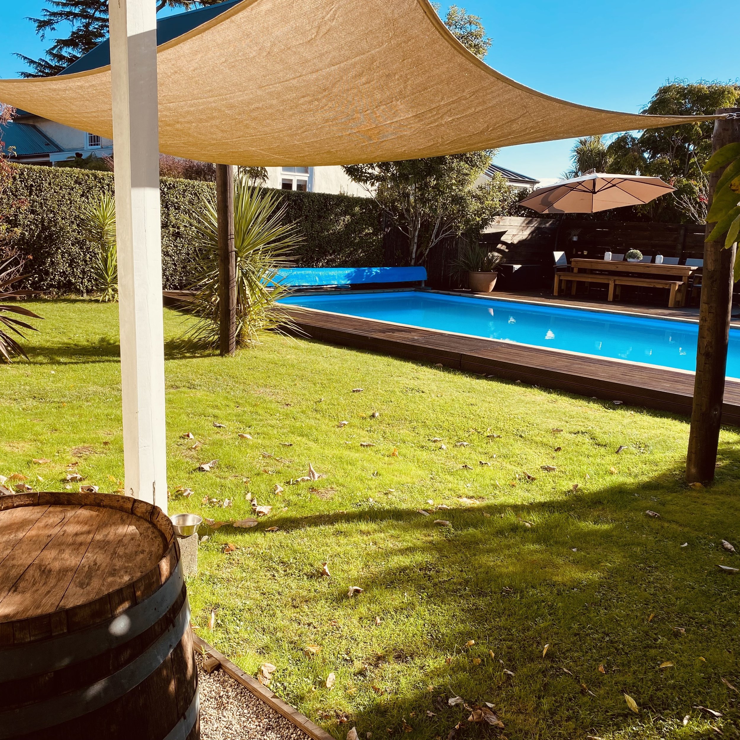 An outdoor backyard scene featuring a swimming pool, a beige shade sail, a beige umbrella, a wooden table, and green plants and trees, with a house visible in the background under a clear blue sky.