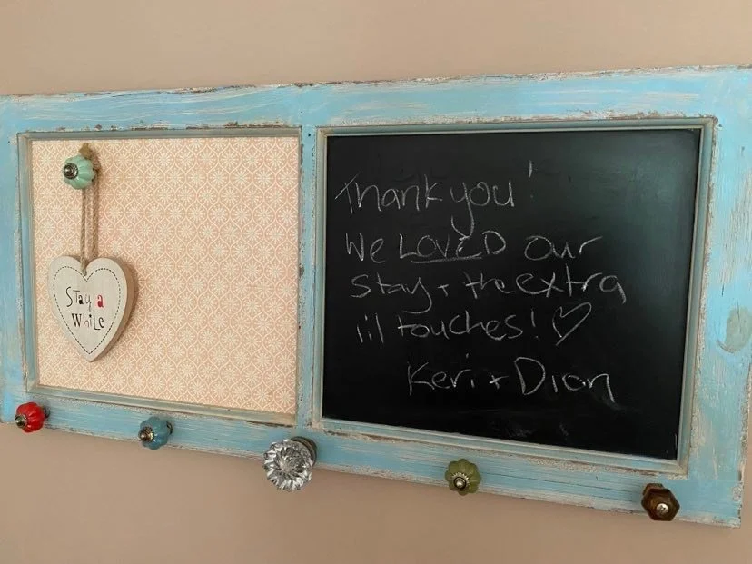 A vintage wall-mounted chalkboard with a handwritten thank you note from Kerr and Dion, and a decorative heart-shaped sign that says 'Stay a While,' attached with colorful knobs.