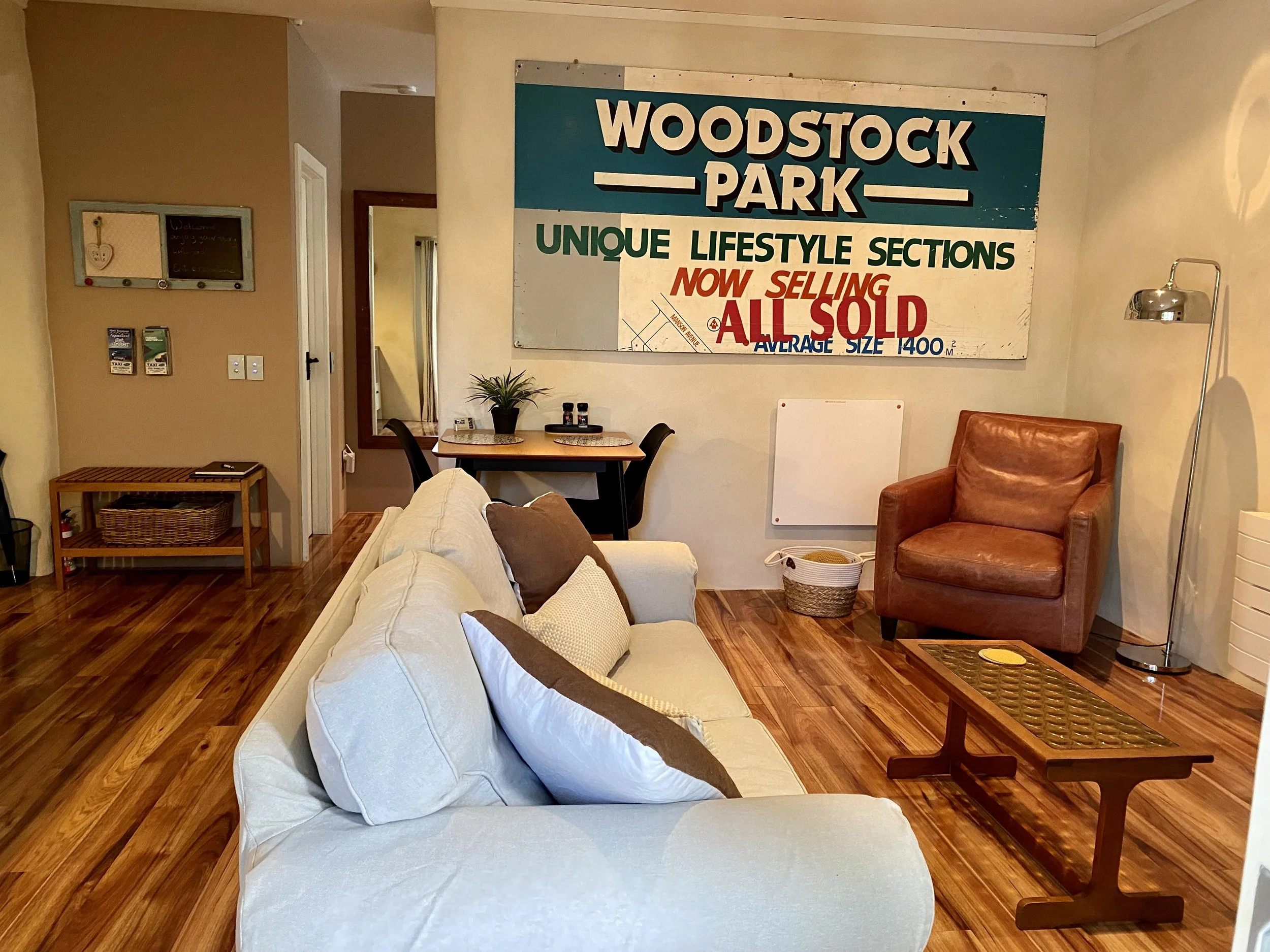 A cozy living room with a white sofa, brown pillows, and a brown leather armchair. A wooden coffee table sits in front of the sofa, and a small table with a basket underneath is on the right. A large wall sign reads 'Woodstock Park, Unique Lifestyle 