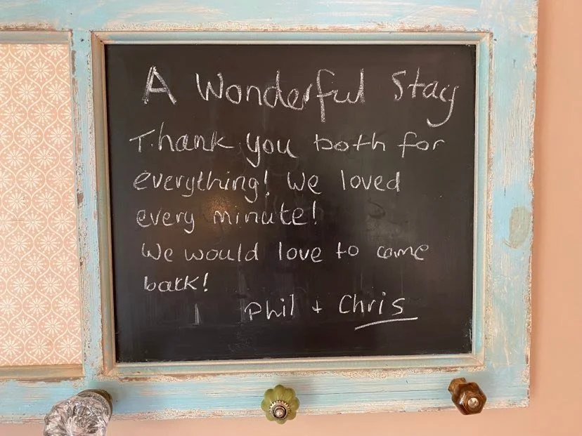 Chalkboard with a thank you message from Phil and Chris at a stay, framed in a weathered light blue frame with decorative wallpaper on the wall.