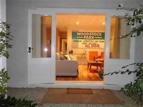 View of a cozy living room seen through an open door, with a white sofa, orange armchair, wooden flooring, and a wall sign that reads 'Woodstock Park' and 'Unique Lifestyle Section'.