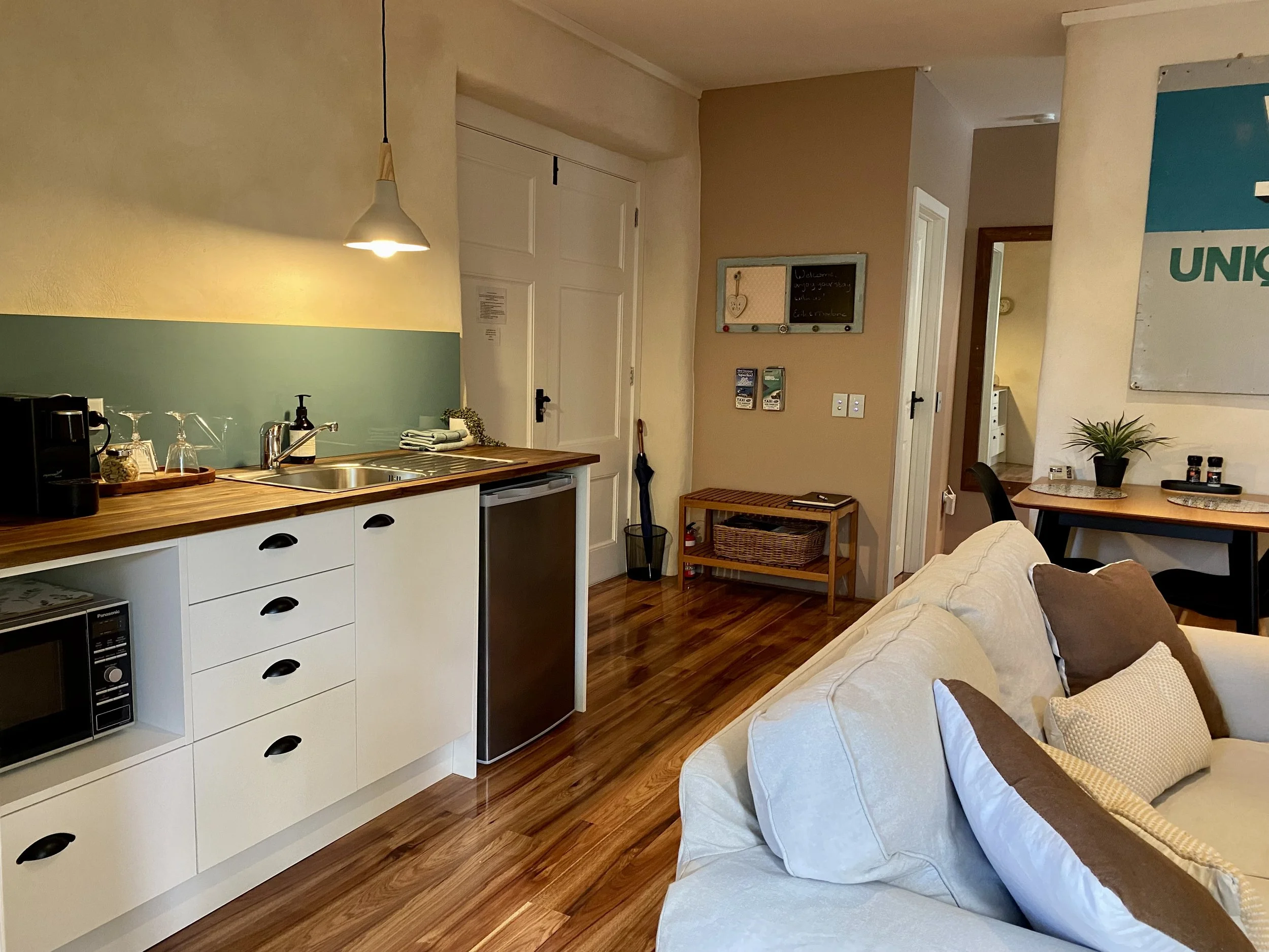 A cozy apartment interior featuring a kitchenette with a white cabinet, black handles, a wooden countertop, a coffee machine, wine glasses, and a dish soap dispenser. Adjacent is a living area with a beige couch, pillows, and a small wooden table. Th