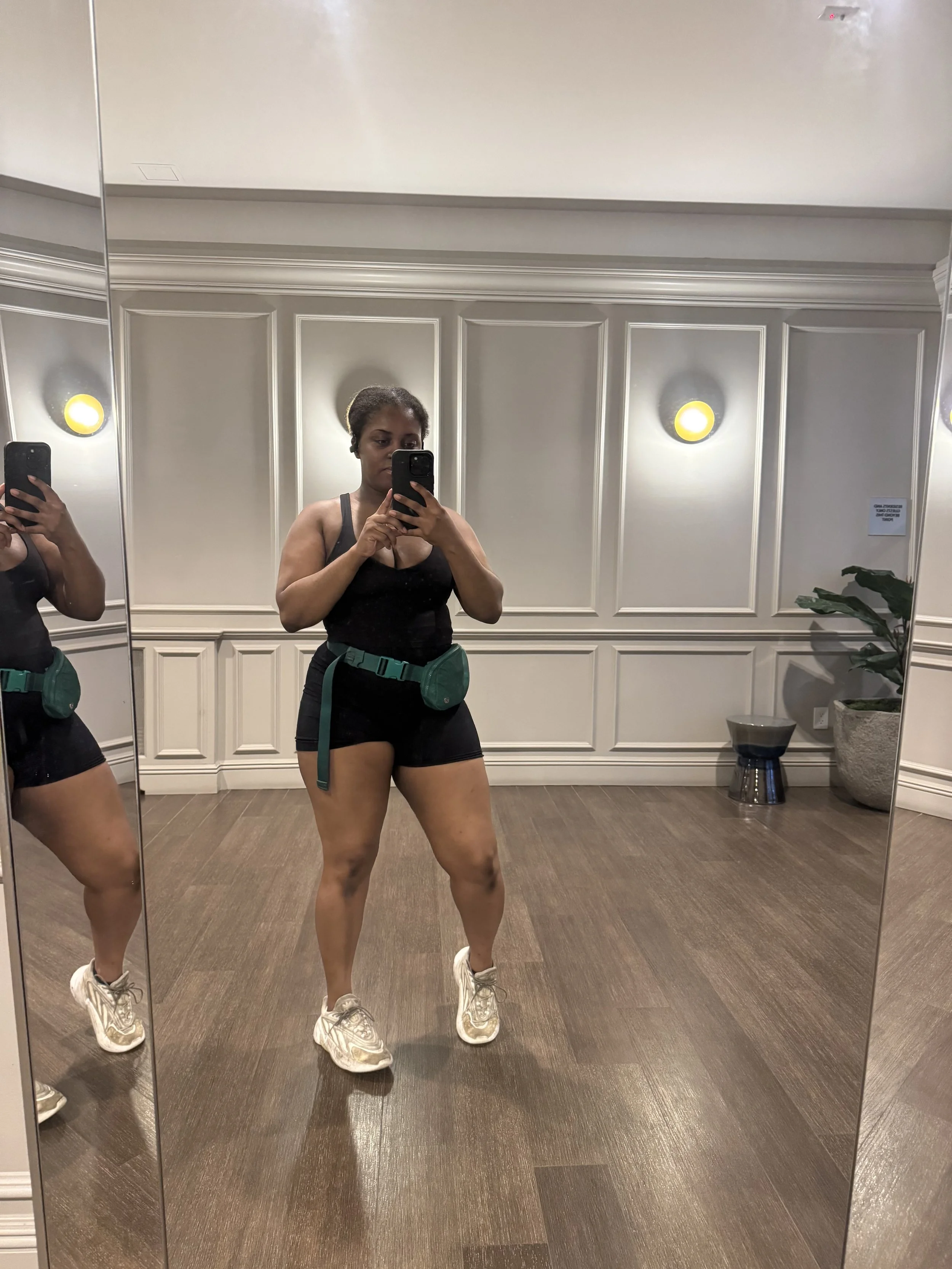 A woman taking a mirror selfie in a well-lit room, wearing a black sports bra, black shorts, and white sneakers, with a green waist bag.