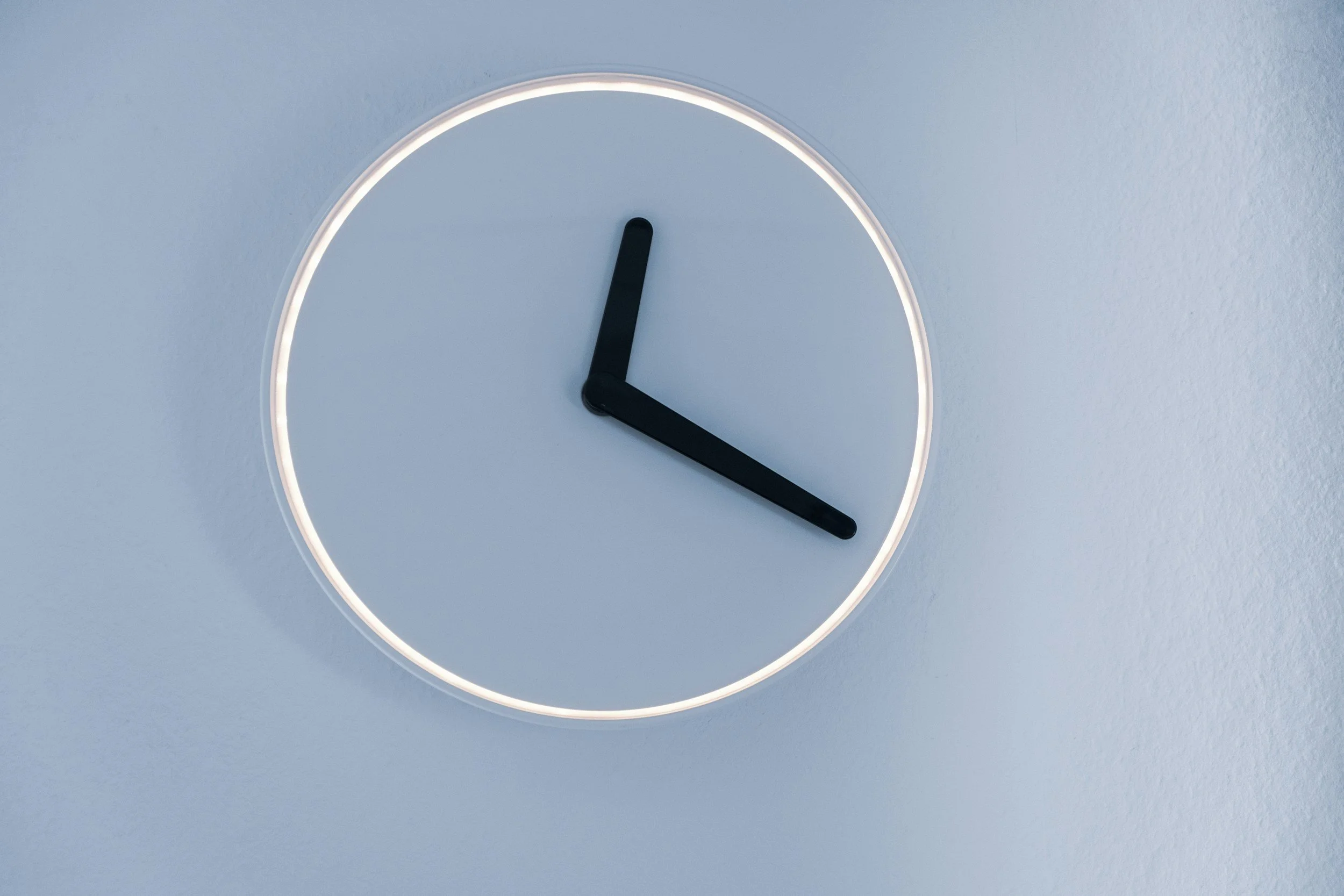 Close-up of a modern, minimalist wall clock with black hour and minute hands against a light grey wall.