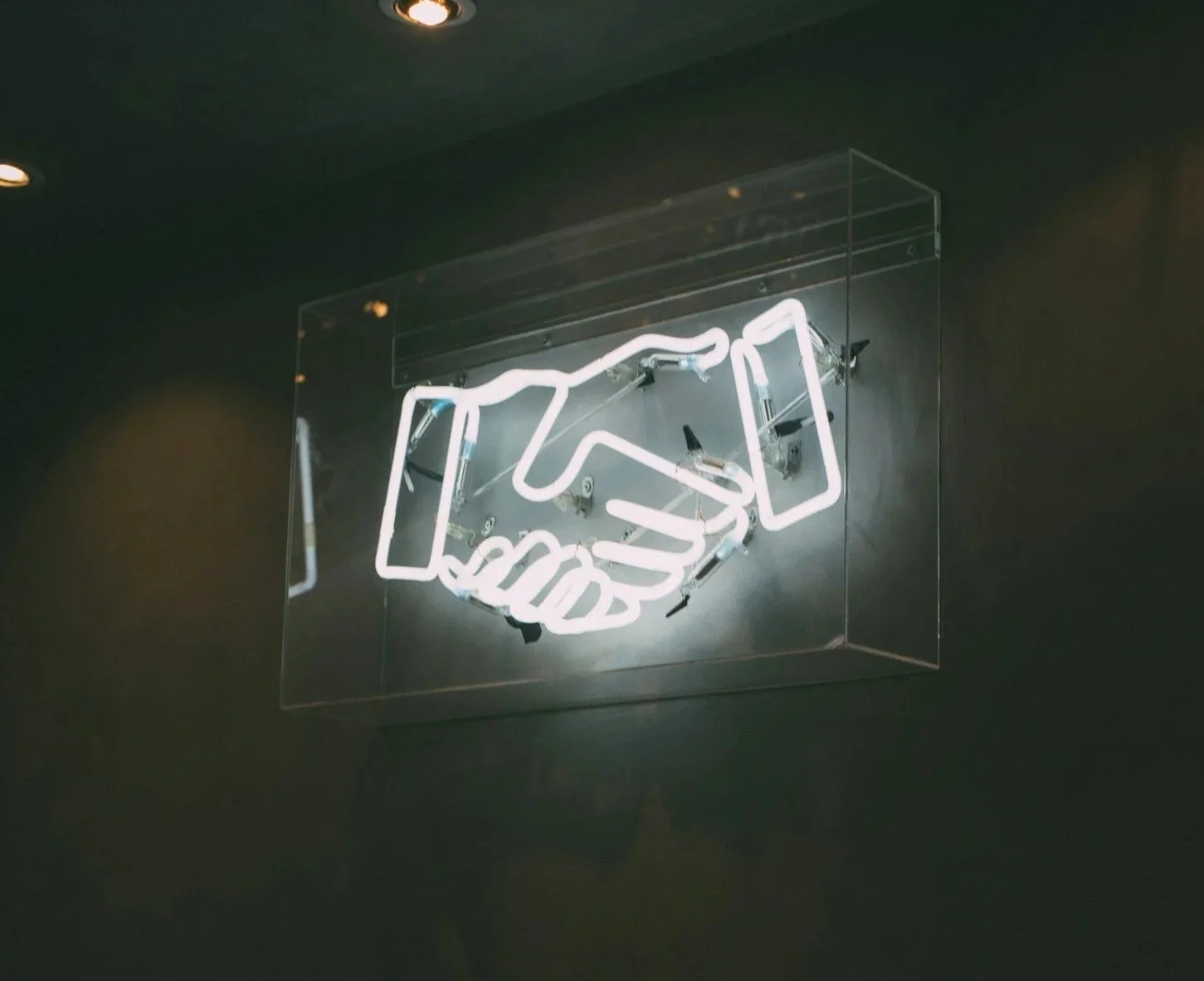 Neon sign with a handshake depicted by two hands in a handshake, mounted on a dark wall.