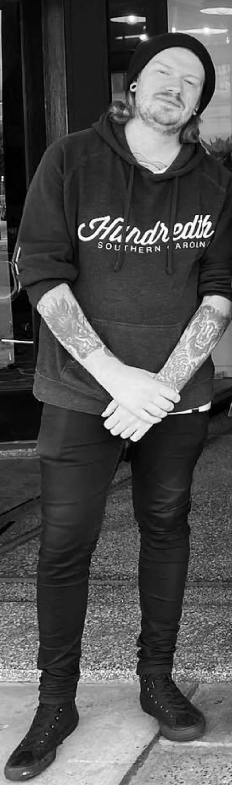 A man with shoulder-length hair, beard, and tattoos on both arms stands outside a building, wearing a beanie, a hoodie that says 'Haddreth Southern Caroline,' and black sneakers, with his hands clasped in front of him.