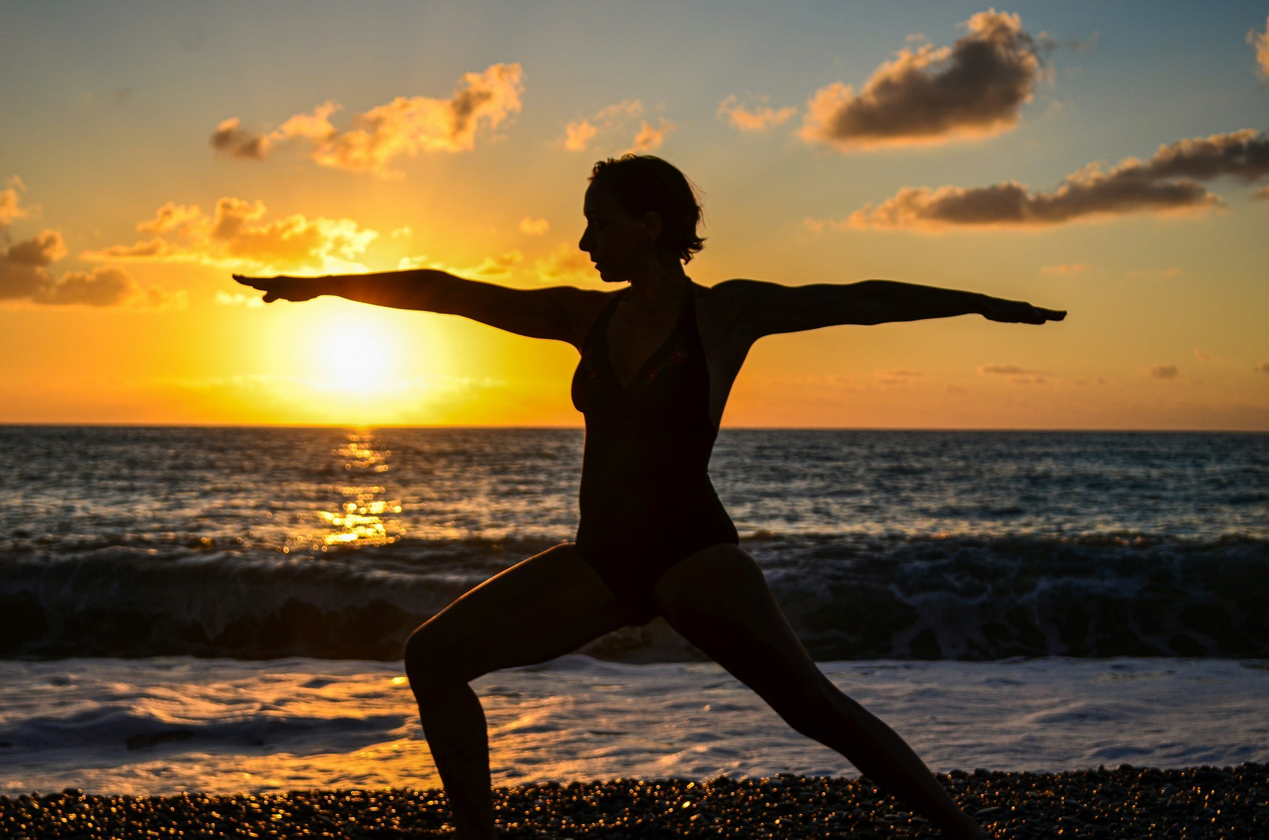 Silhouette of a woman practicing yoga on the beach during sunset with clouds in the sky and the ocean in the background.