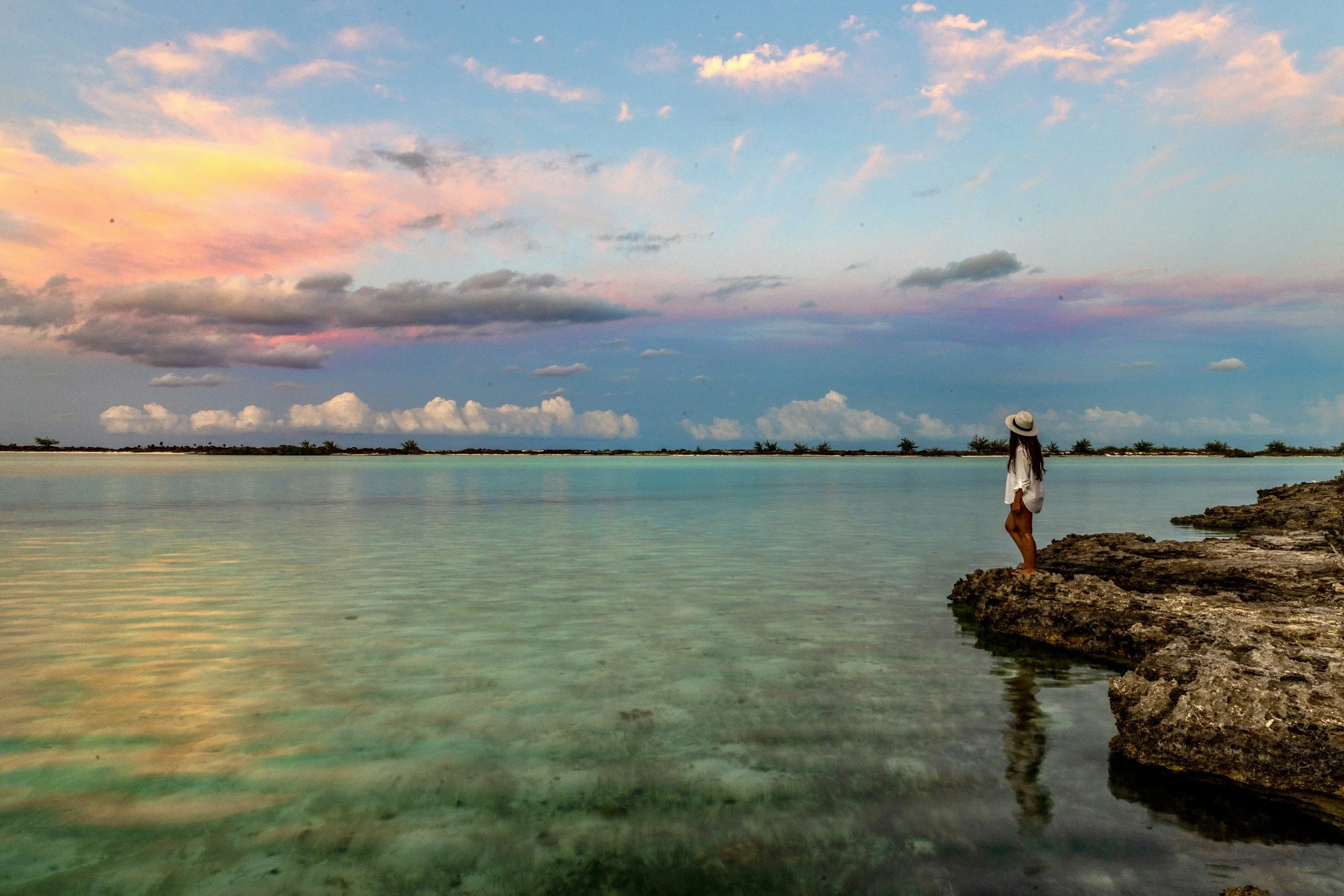 A woman with long hair wearing a hat and white shirt standing on rocks by the water during sunset or sunrise, with a sky filled with pink, orange, and blue clouds.