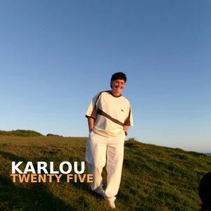Smiling young man standing outdoors on a grassy hill under a clear blue sky, wearing a white outfit with dark accents, with the caption 'KARLOU TWENTY FIVE'.