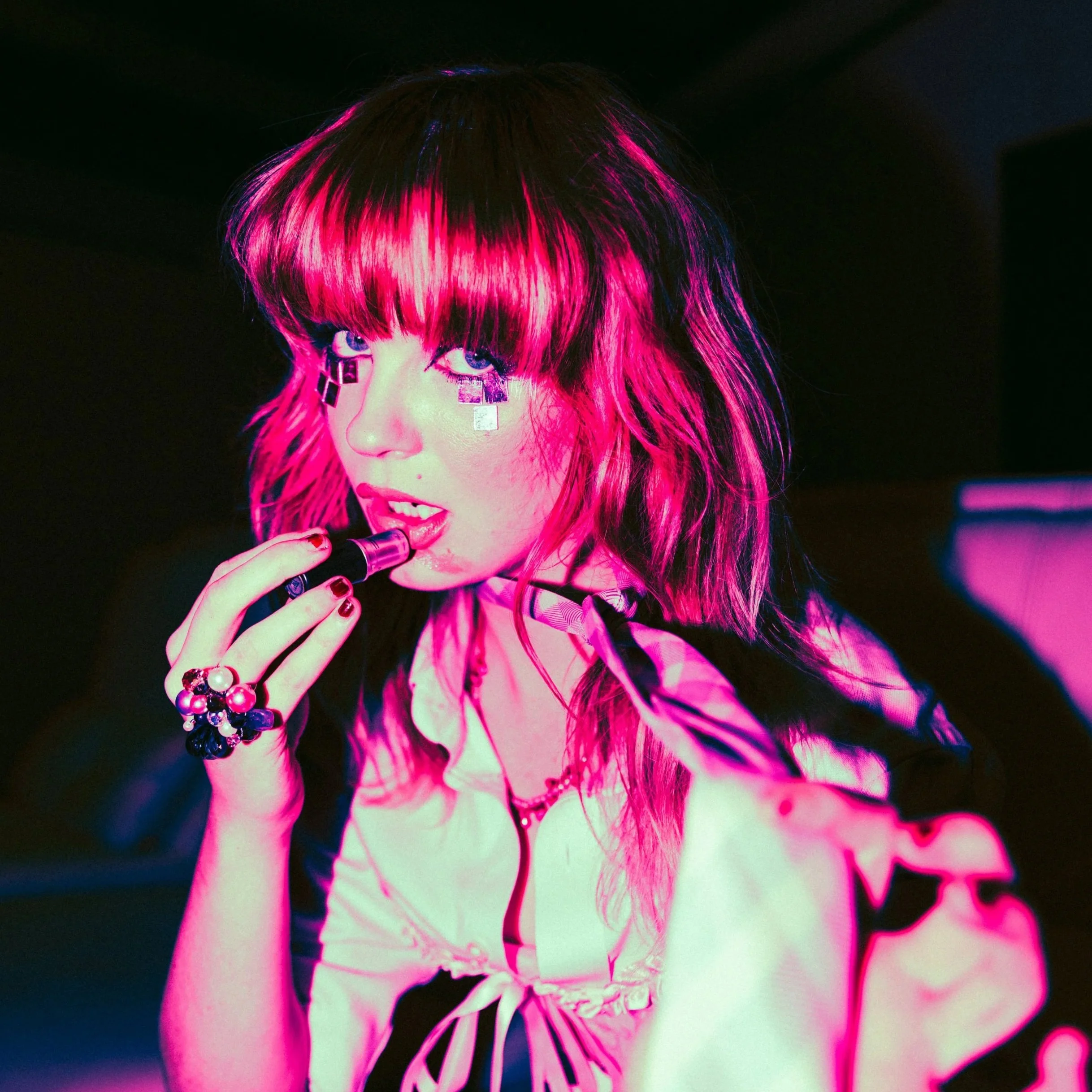Close-up of a person with vibrant pink and black hair, wearing bold makeup, jewelry, and colorful clothing, with a dark background.