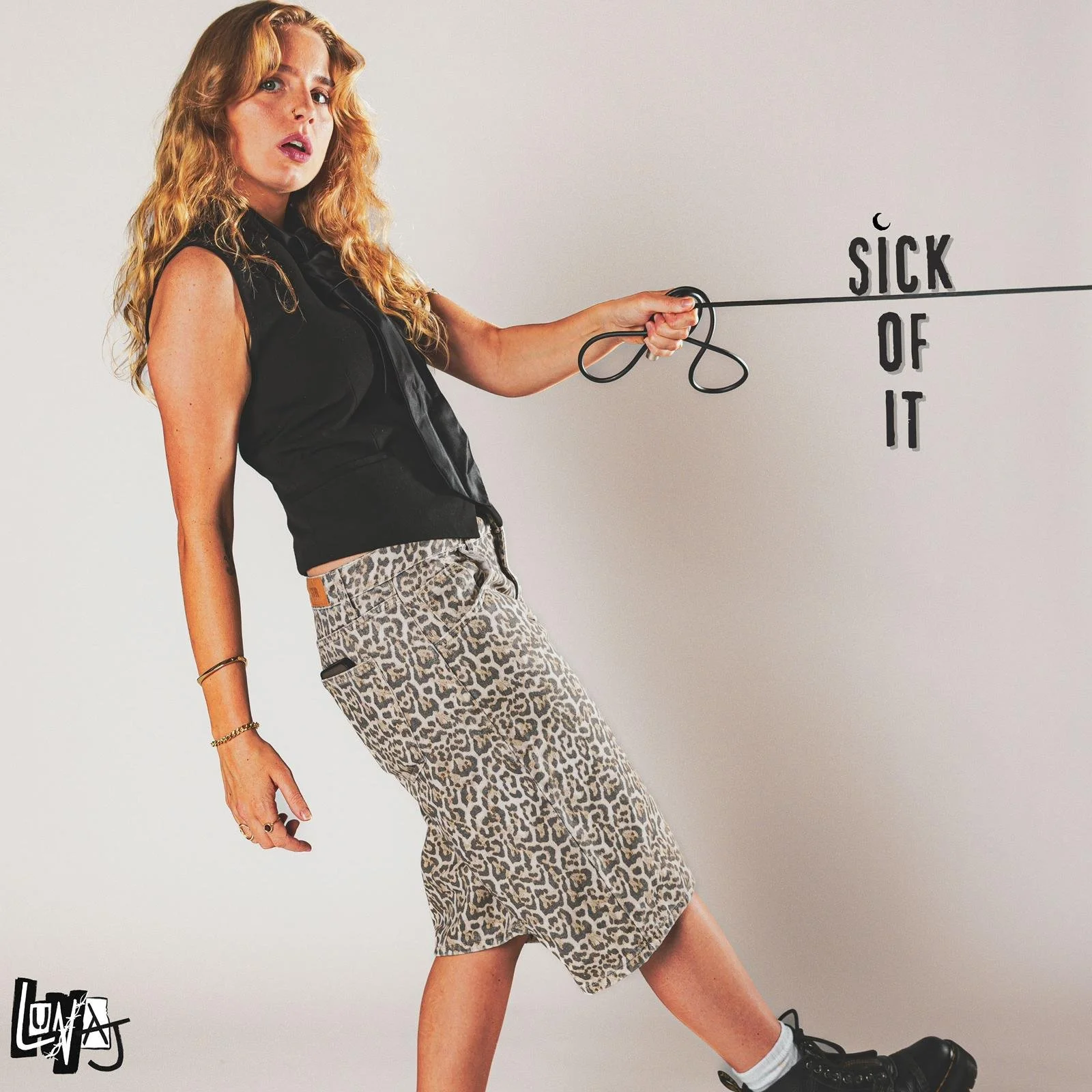 A woman with wavy blonde hair wearing a black sleeveless top, leopard-print shorts, and black boots, holding a stick that points to the words 'SICK OF IT' on a plain background.