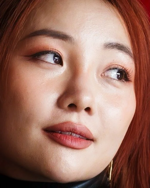 Close-up of a woman with red hair looking to the side, wearing makeup including eyeshadow and lipstick.
