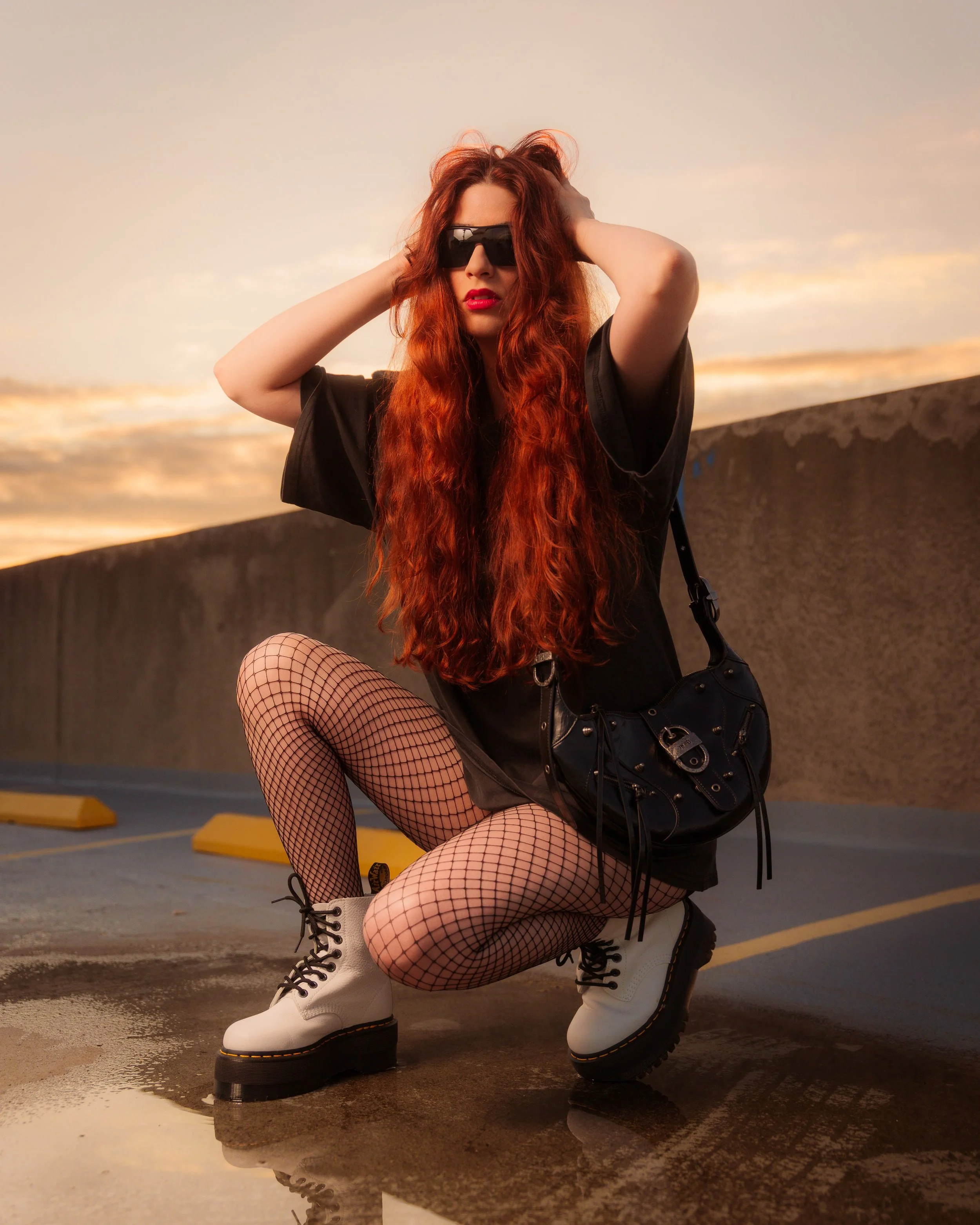 A woman with long red hair wearing black sunglasses, red lipstick, a black shirt, fishnet stockings, white combat boots, and a black leather bag in a parking lot at sunset.
