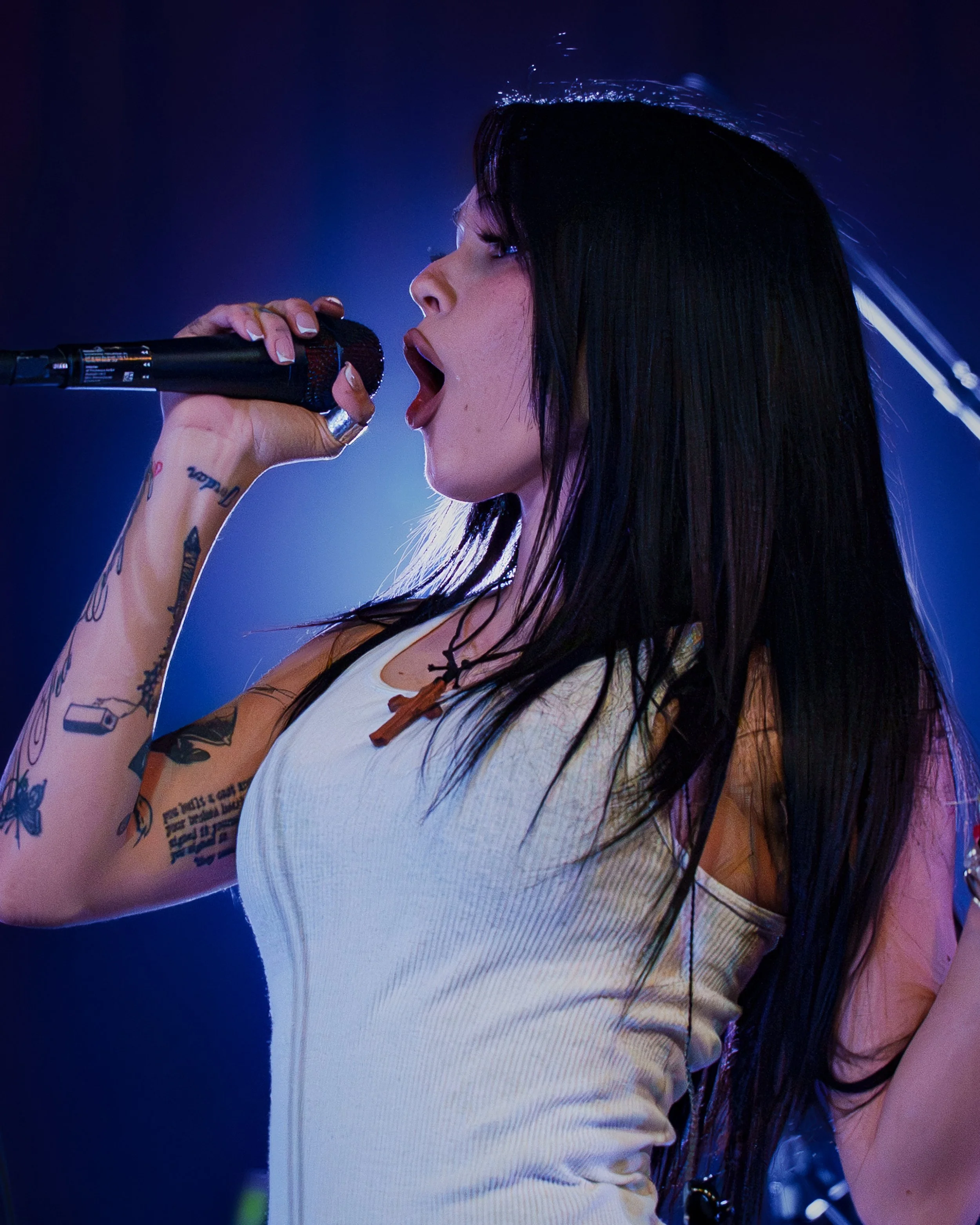 A woman with long, wet black hair singing into a microphone on stage, wearing a sleeveless beige top, with tattoos on her arm, under blue lighting.
