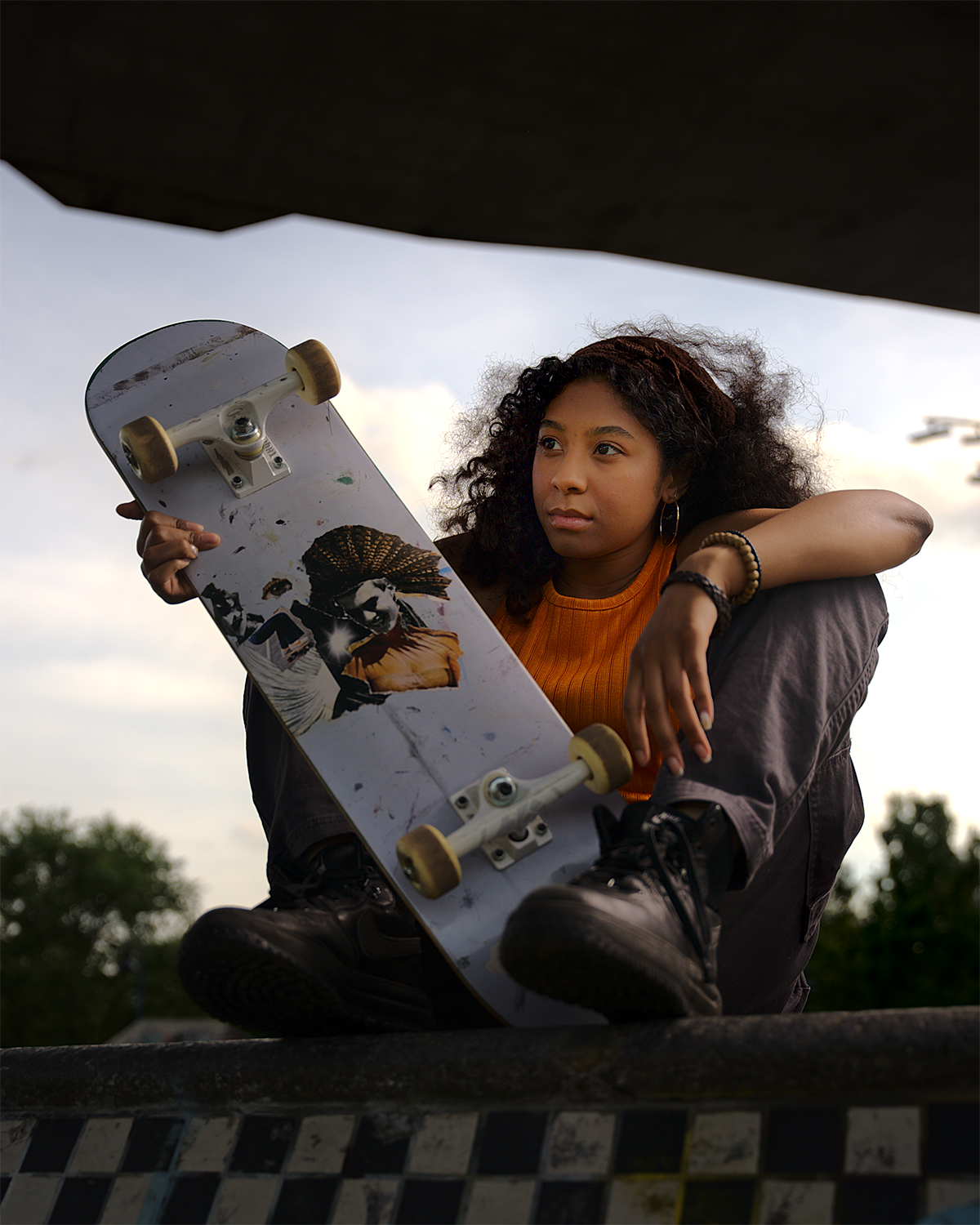 A young woman sits under a skate park, holding a skateboard with a graphic of a woman in a feathered headdress on it. She has curly hair, hoop earrings, and is wearing an orange shirt and gray pants, with a serious expression.