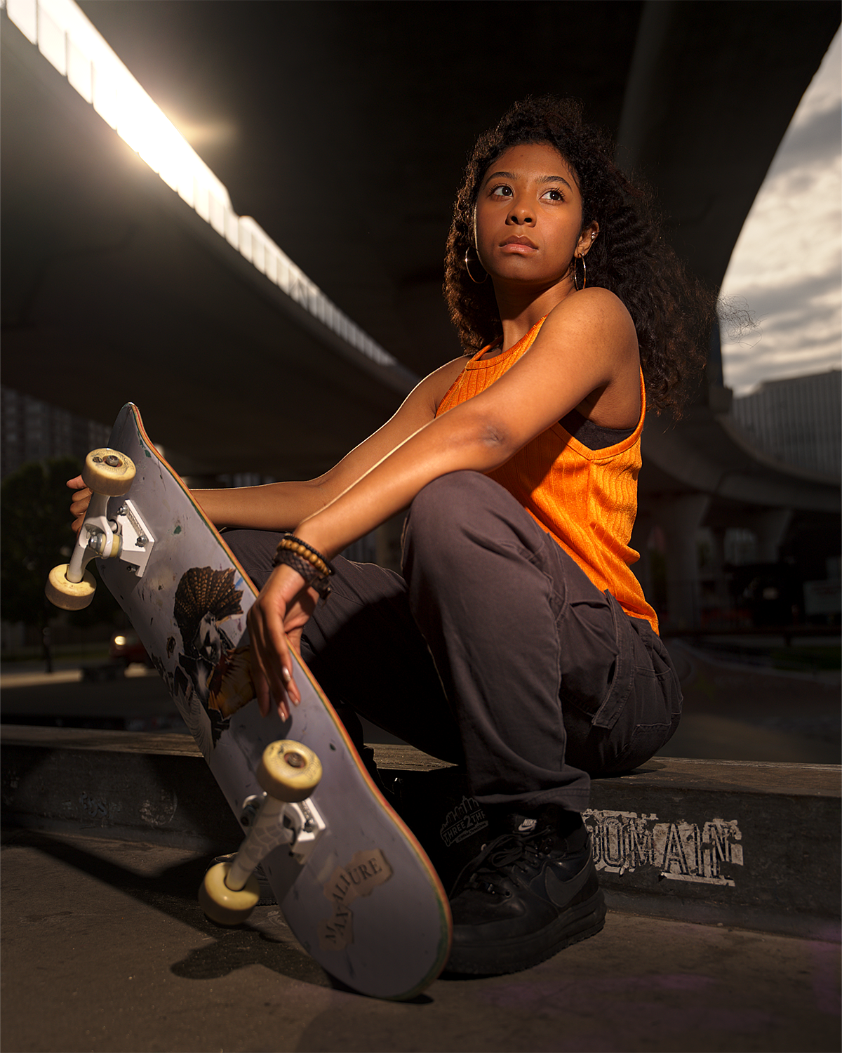 A young woman with curly hair, wearing an orange sleeveless top and black pants, is sitting on the ground under a bridge with a skateboard beside her. The sky is partly cloudy, and there are buildings and a road in the background.