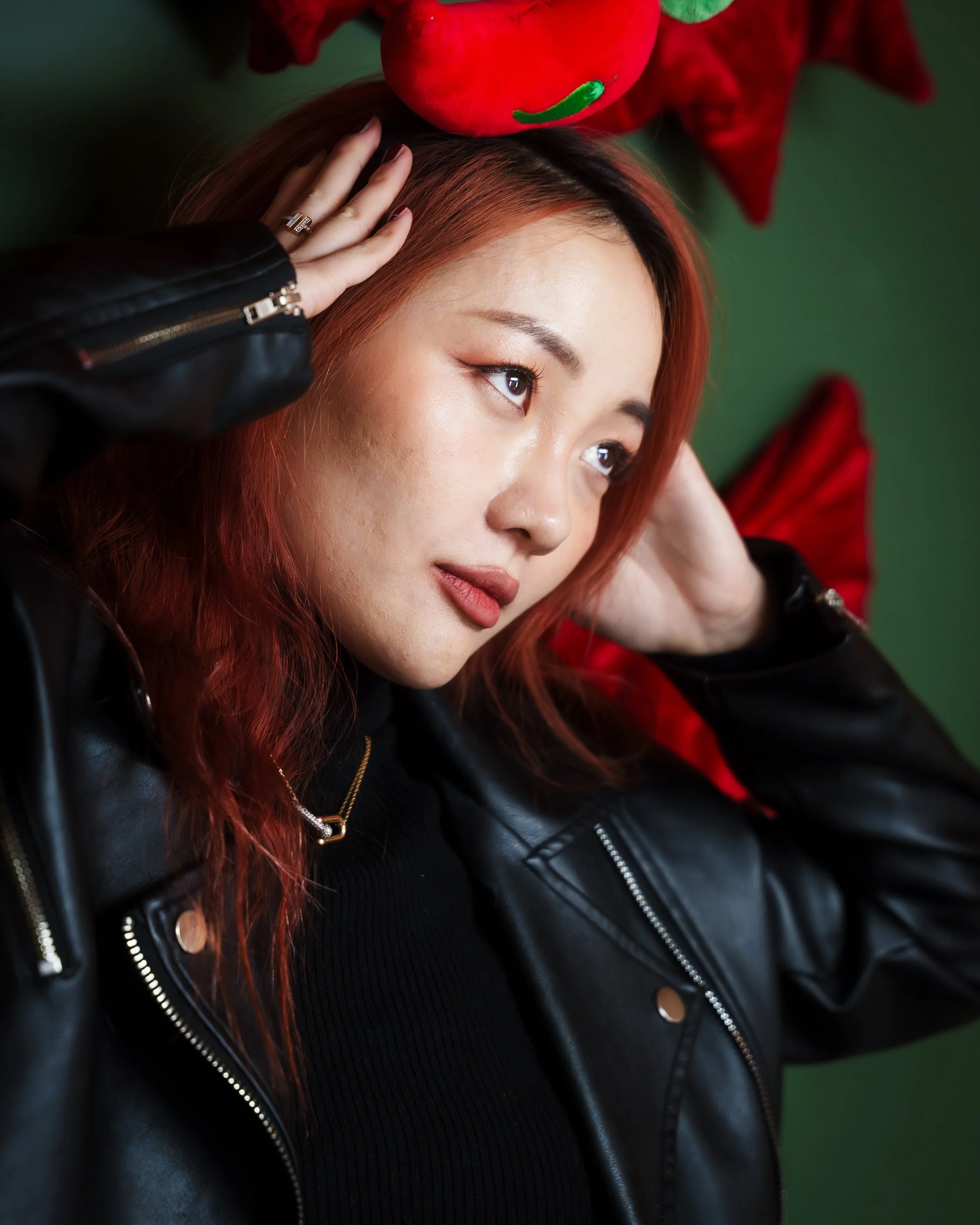 A woman with red hair wearing a black leather jacket, holding her head with both hands, and a large red flower on her head, against a dark green background.