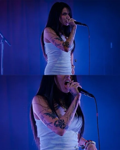 Female singer with long dark hair, tattoos, and a white tank top performing on stage with a microphone, illuminated by blue lighting.