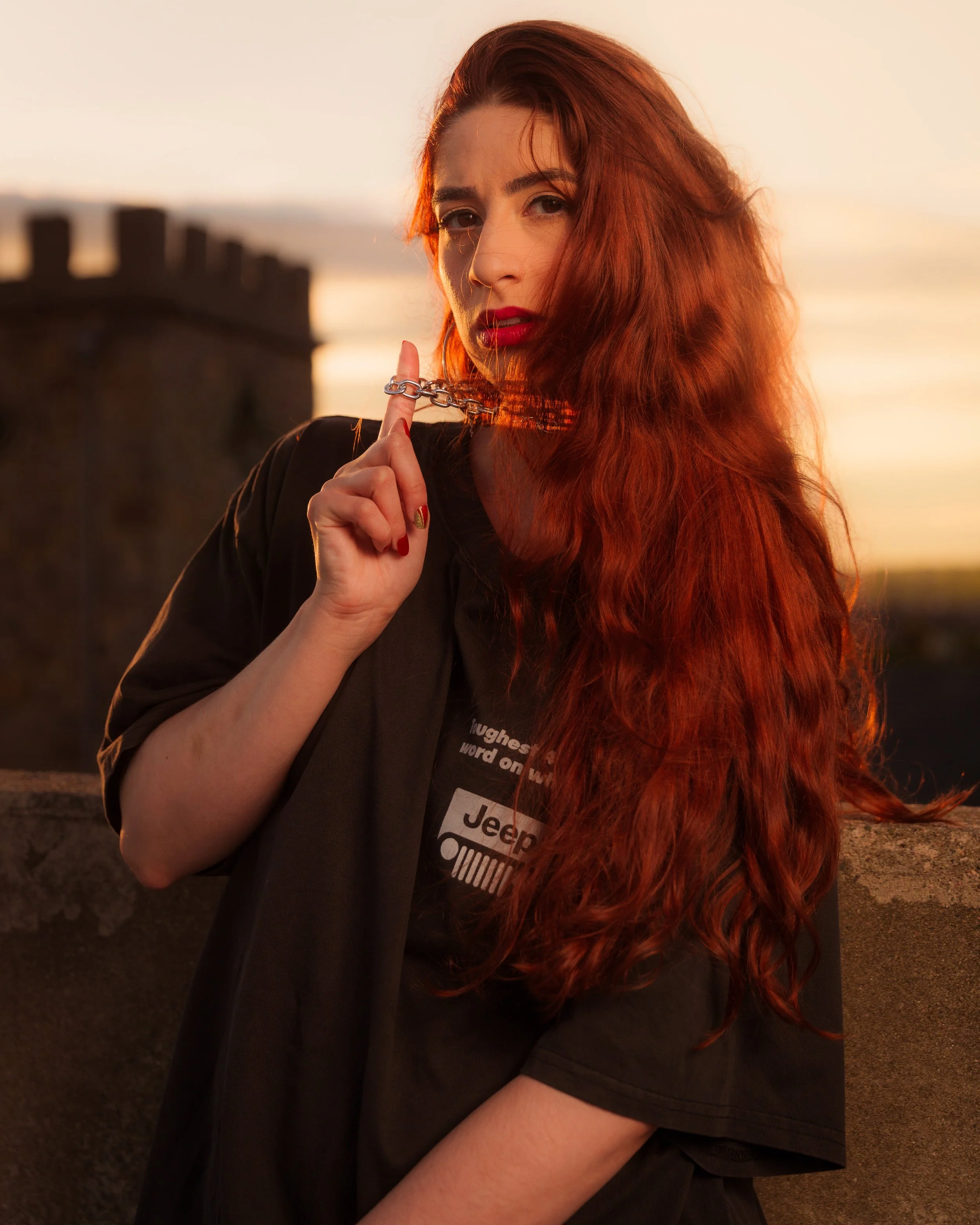 A woman with long red hair and red lipstick holds a silver chain near her lips. She is wearing a black shirt with a Hyundai logo and a name tag. The background features a sunset with warm tones.