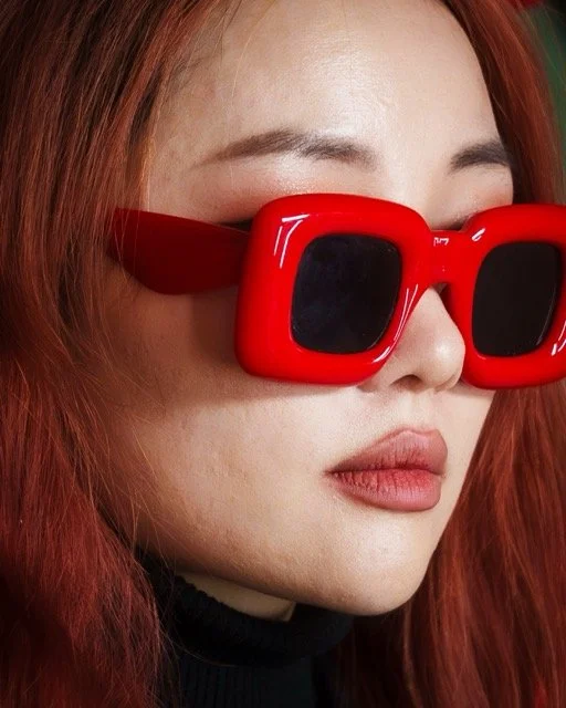 Close-up of a woman wearing large red sunglasses with dark lenses and red hair.