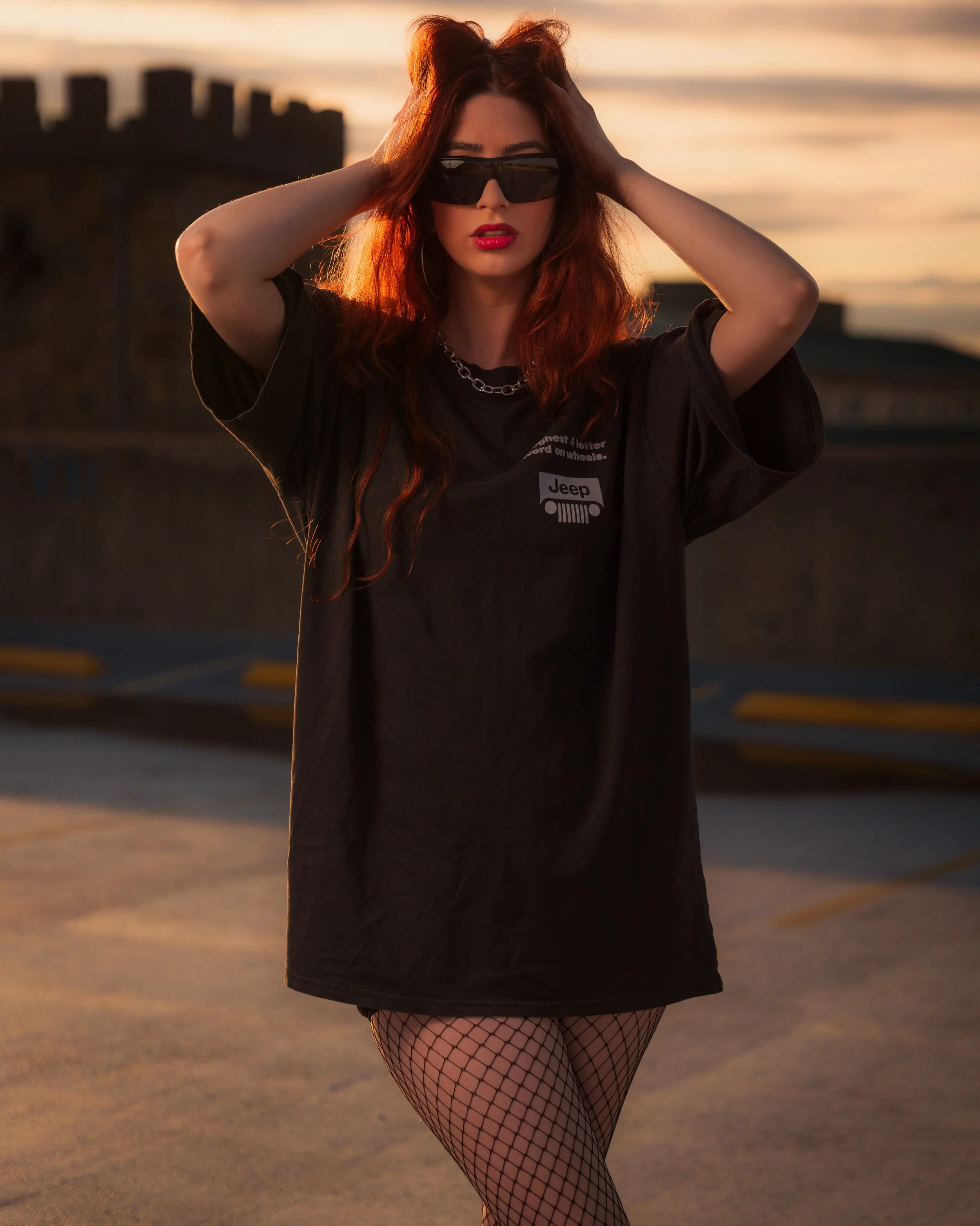 A woman with long red hair wearing sunglasses, a black oversized T-shirt with a Jeep logo, fishnet stockings, and earrings, standing outdoors during sunset with her hands on her head and a blurred background of a parking lot and buildings.