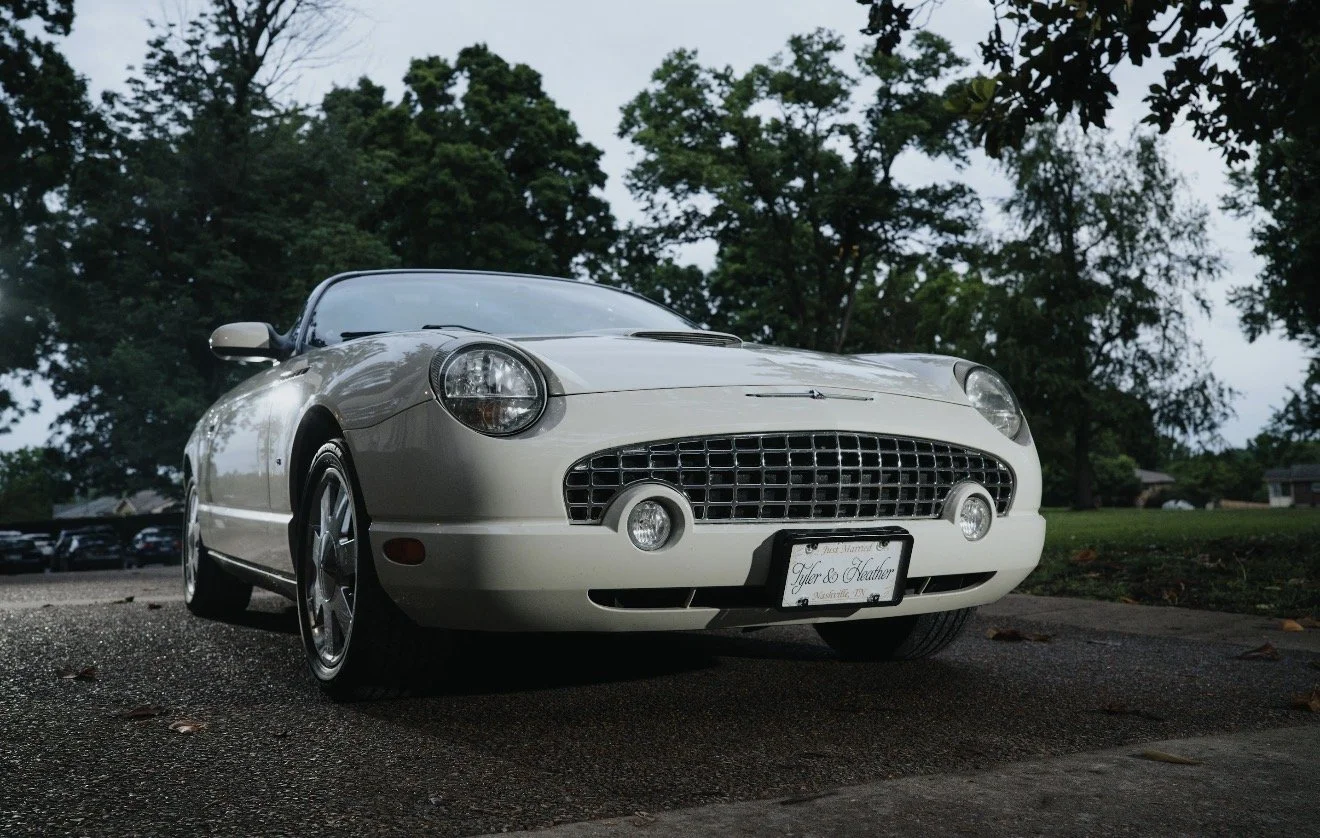 A white vintage-style sports car parked on a driveway with trees in the background, taken during dusk or early evening.
