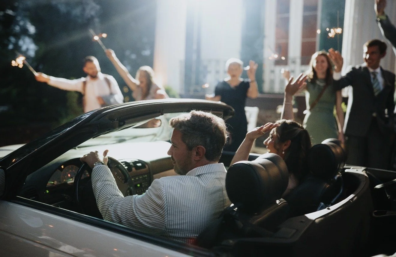 People celebrating a wedding with a newlywed couple in a convertible car, guests holding sparklers in the background.