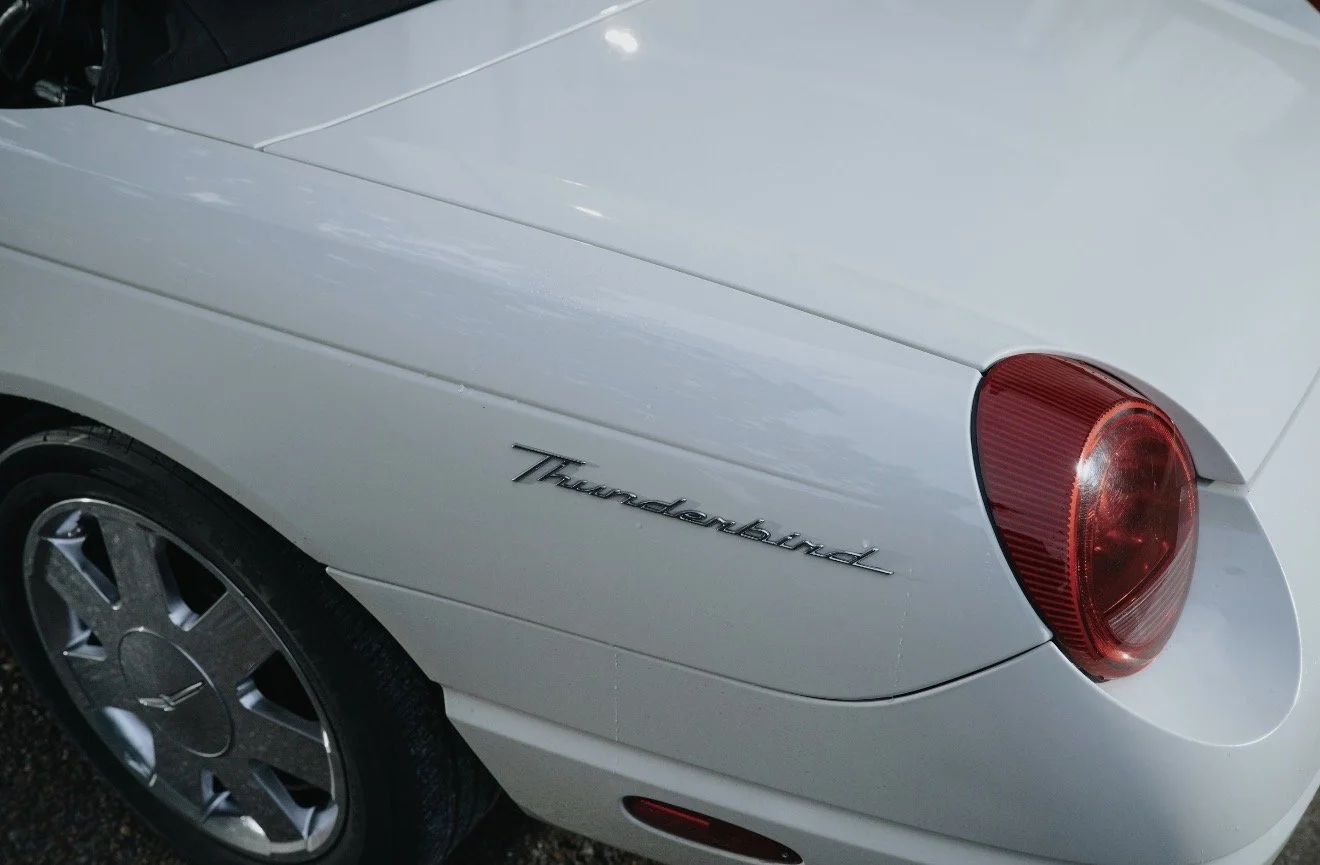 Close-up of a white car's rear section, showcasing the 'Thunderbird' emblem and a red circular tail light.