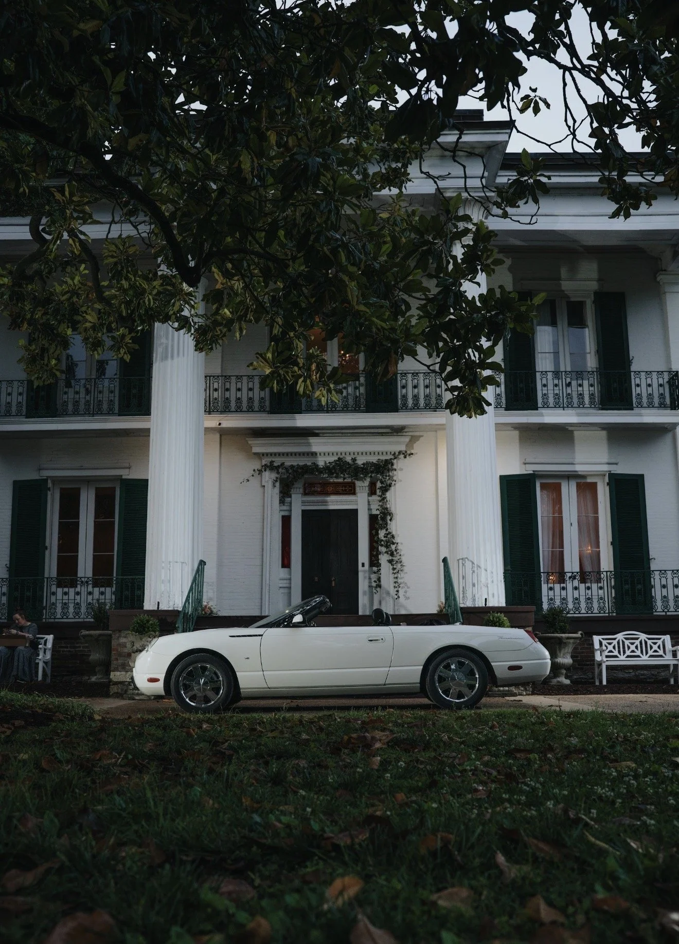 Front view of a white convertible sports car parked in front of a large white house with tall columns and green shutters, trees partially obscuring the house, and a bench on the right side.
