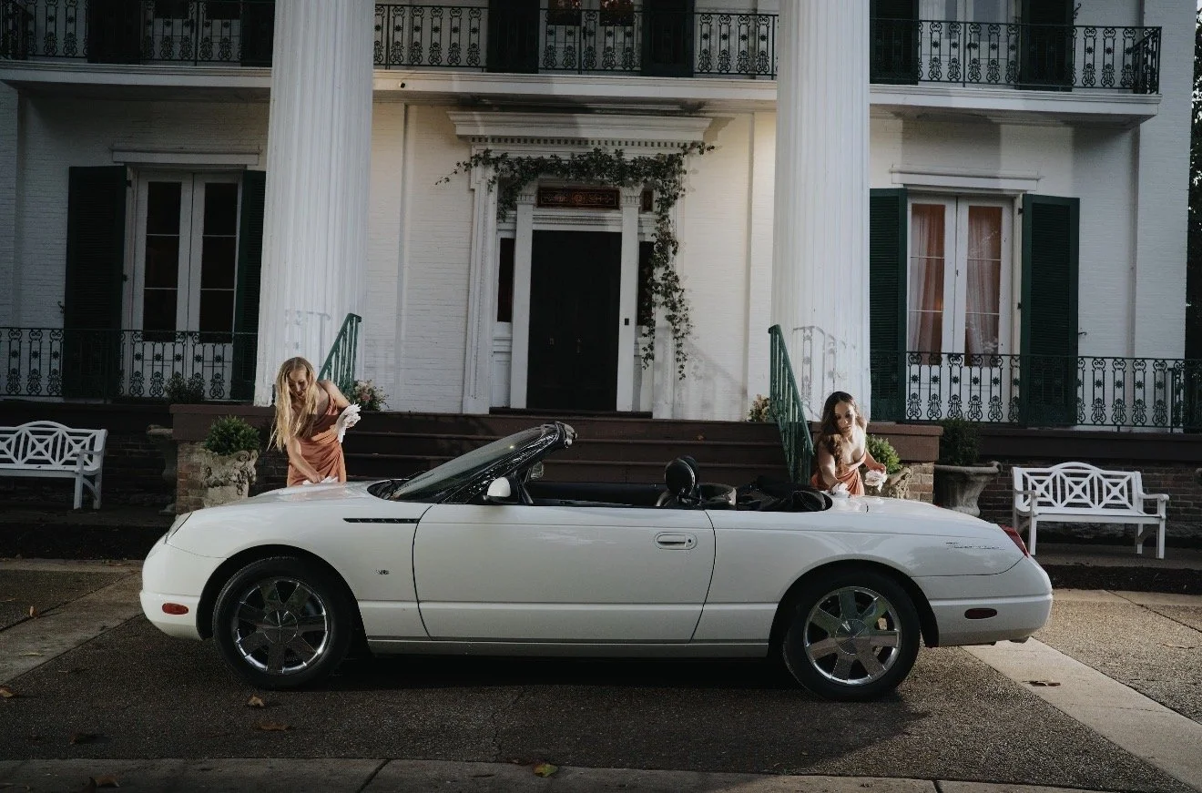 A white convertible car parked in front of a large house with white columns. Two women in peach dresses are standing by the car, one on each side, near the house's steps.