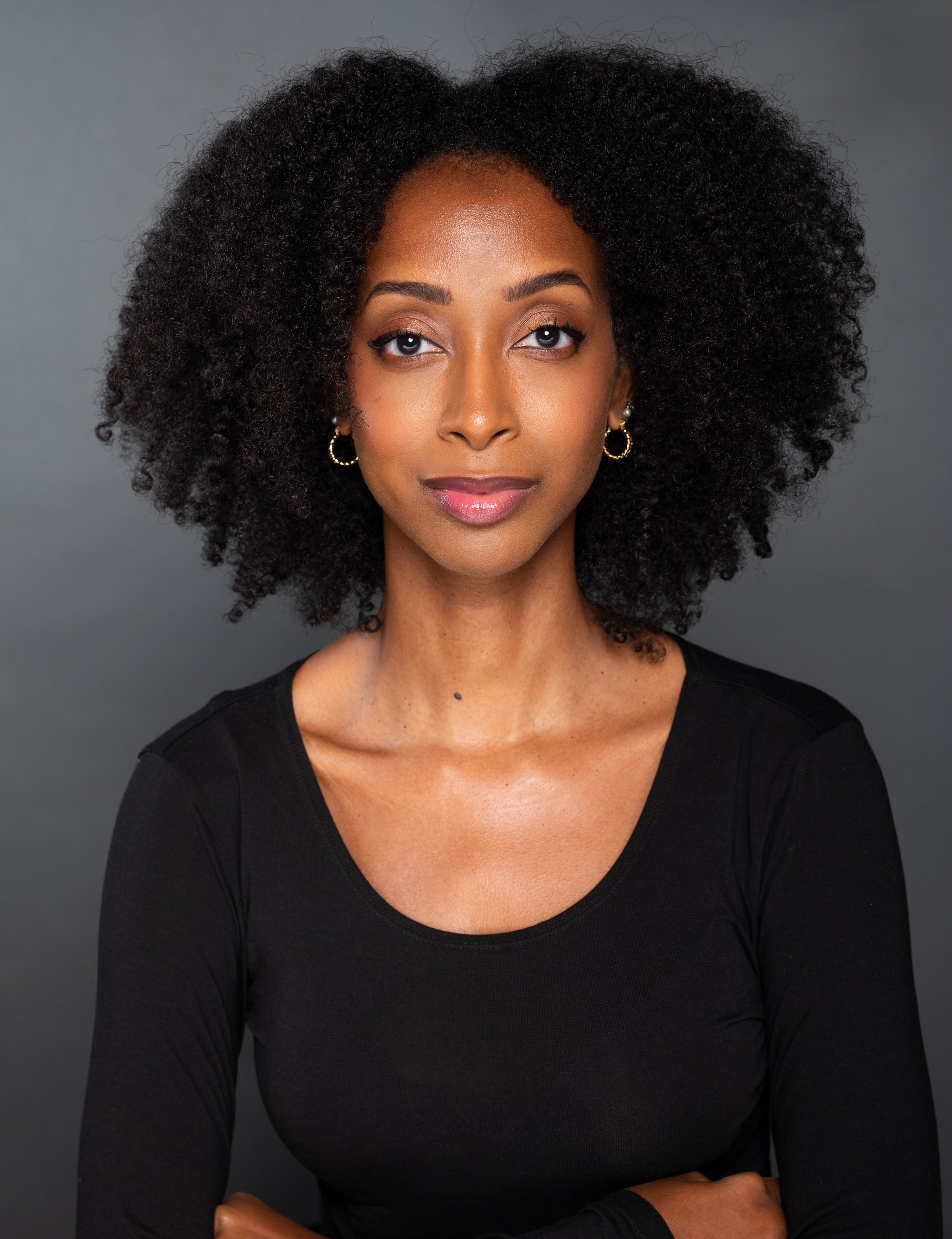 A portrait of a woman with dark skin and curly black hair wearing a black top, gold earrings, and makeup with natural lips and eyeshadow, posing against a plain, dark gray background.