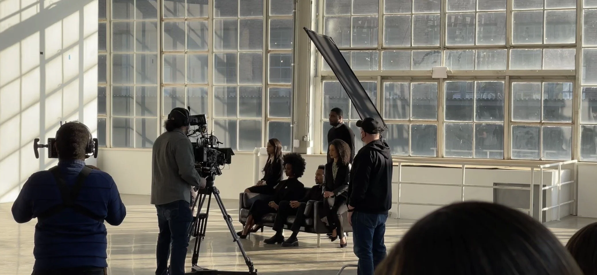 A film crew is filming a scene inside a bright industrial-style space with large windows. Several actors are seated on a sofa and standing, while crew members operate cameras and lighting equipment.