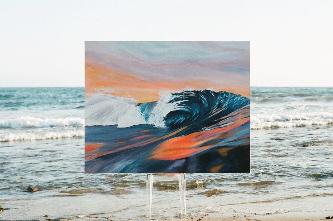A painting of a large ocean wave with a sunset sky in the background, placed on a clear stand at the beach.