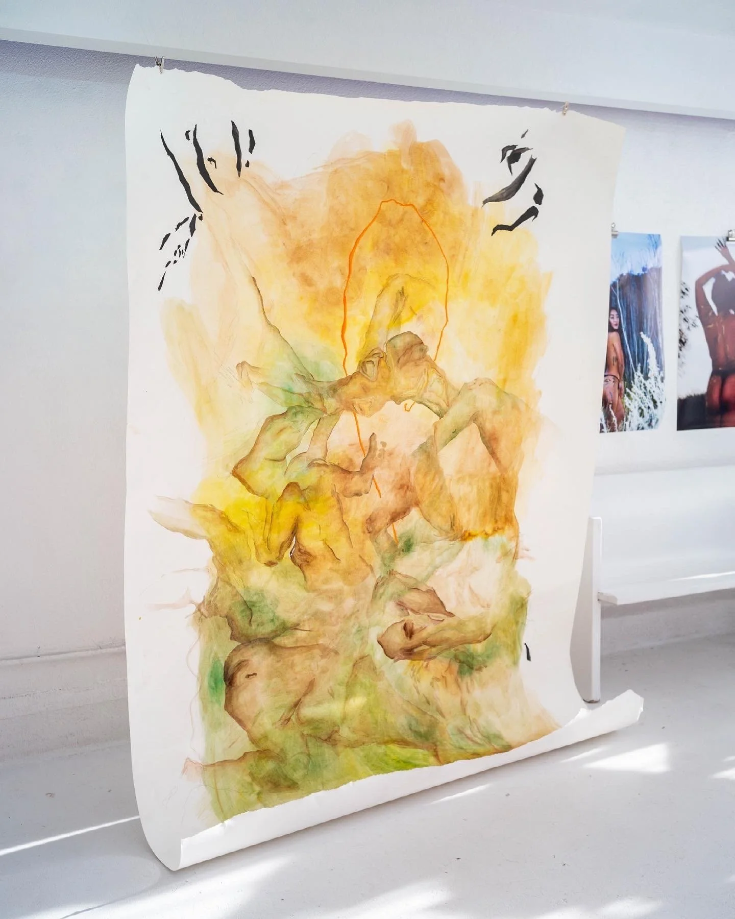 An abstract watercolor artwork on paper featuring earthy tones of yellow, orange, brown, and green with sparse black strokes, displayed on a white wall with other art pieces in the background.