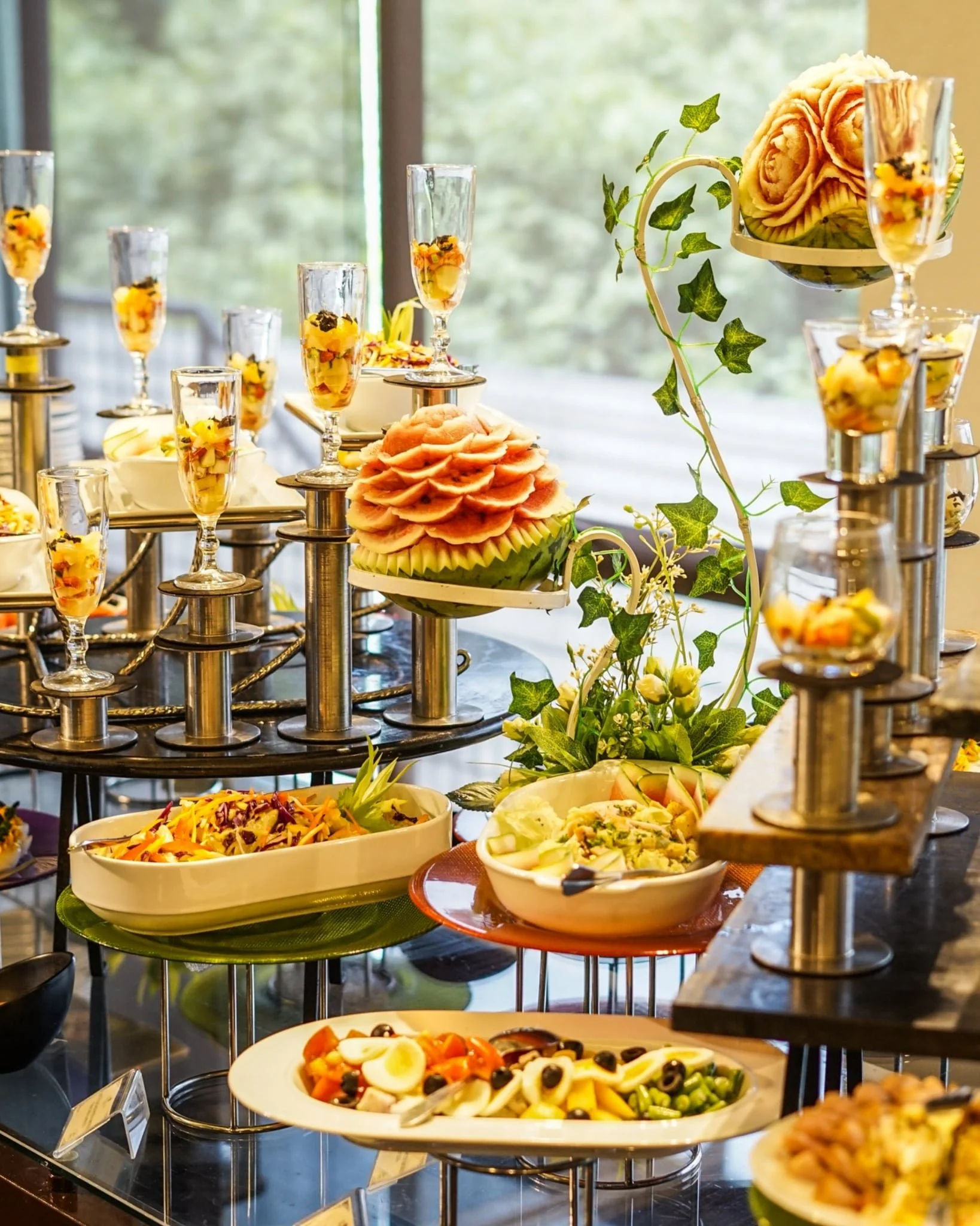 A buffet table with various dishes, including salads, fruit, and sliced vegetables, decorated with greenery and surrounded by tall glass containers.