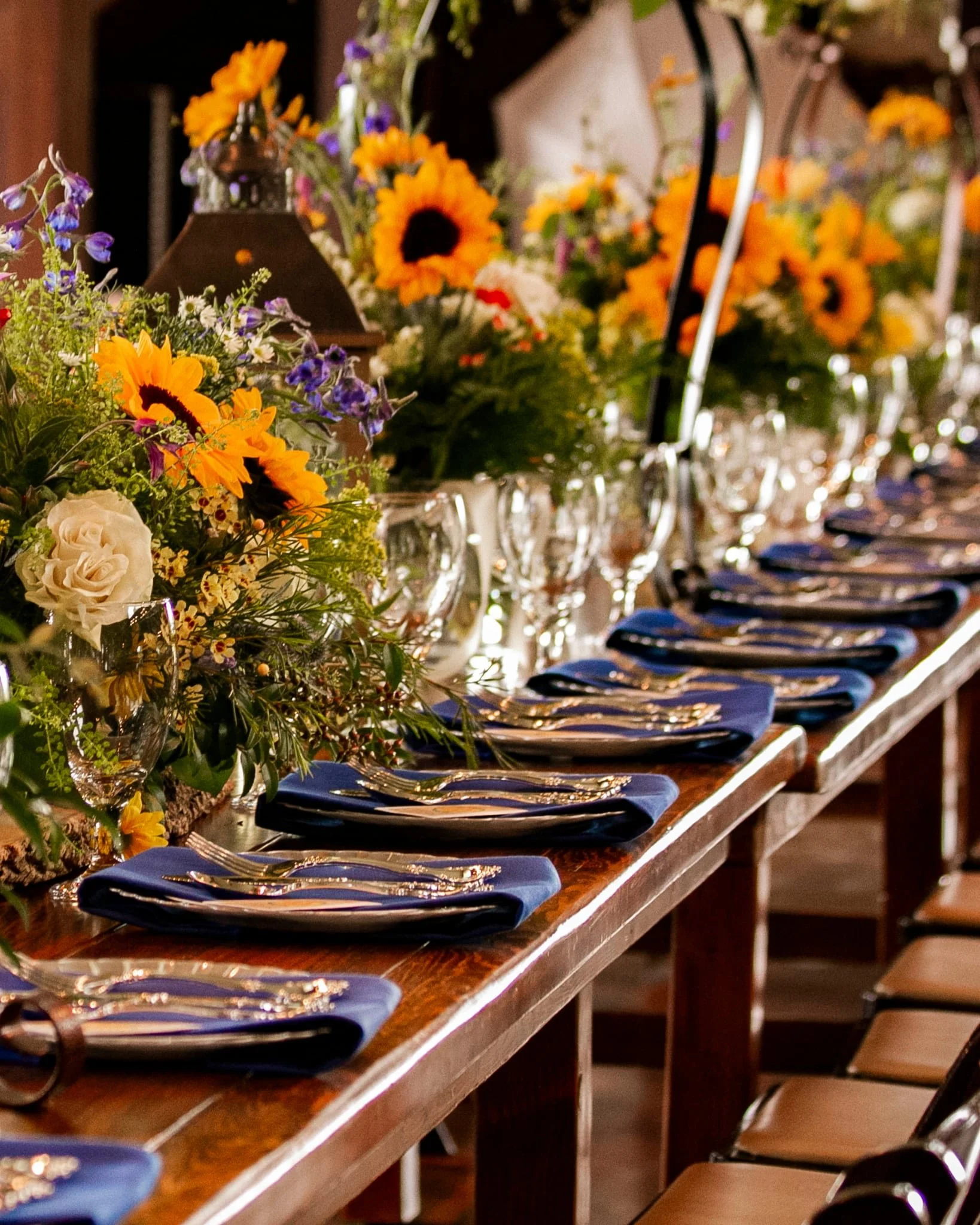 A long dining table set for a banquet, decorated with vibrant floral arrangements, gold and blue dinnerware, and blue cloth napkins, in a warmly lit room.