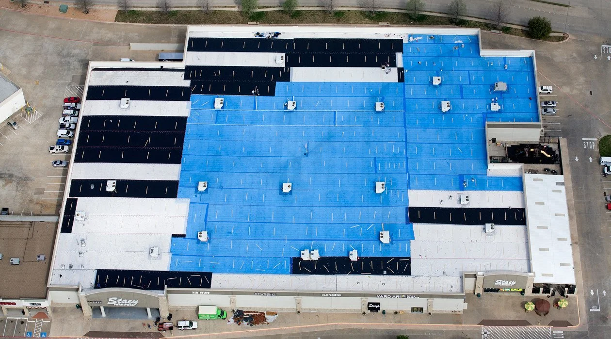 A large rooftop with sections of black and blue roofing, with several HVAC units installed. The building is surrounded by a parking lot with cars and a sidewalk with pedestrians.