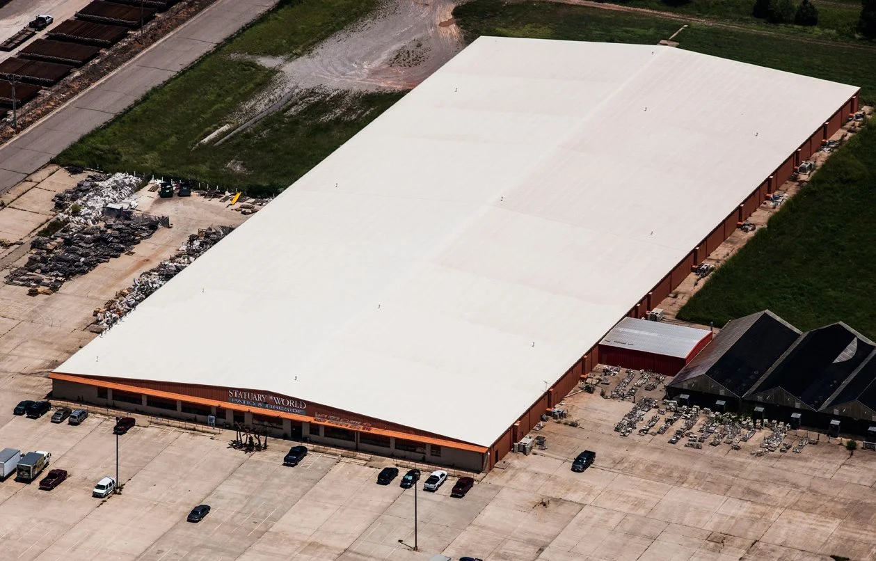 Aerial view of a large building with a white roof, parking lot with cars in front, and surrounding areas with equipment and green spaces.