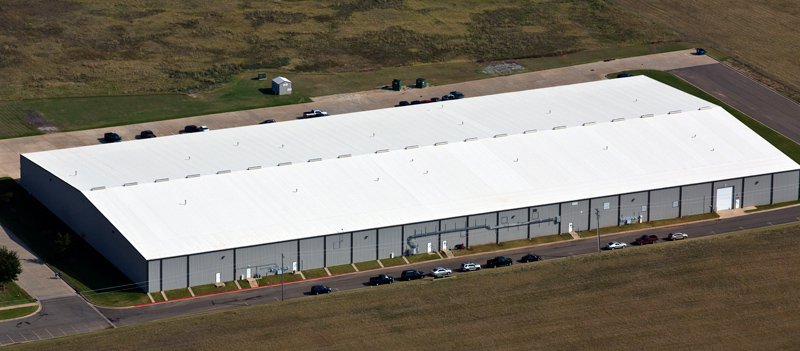 Large industrial warehouse with a white roof and gray exterior, surrounded by parking lot and grassy fields.