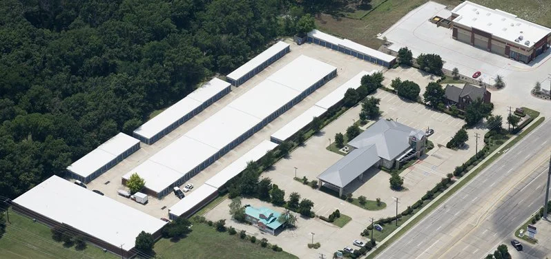 An aerial view of a large event tent area with multiple white tents, a parking lot, a small building, and a pool with a palm tree design, surrounded by trees and roads.