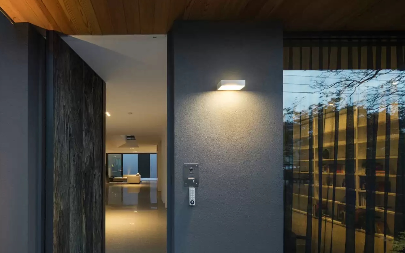 View of the entrance to a modern house showing a wall-mounted light and intercom, with an interior hallway leading to a living room with a sofa, and large windows reflecting bare trees outside.