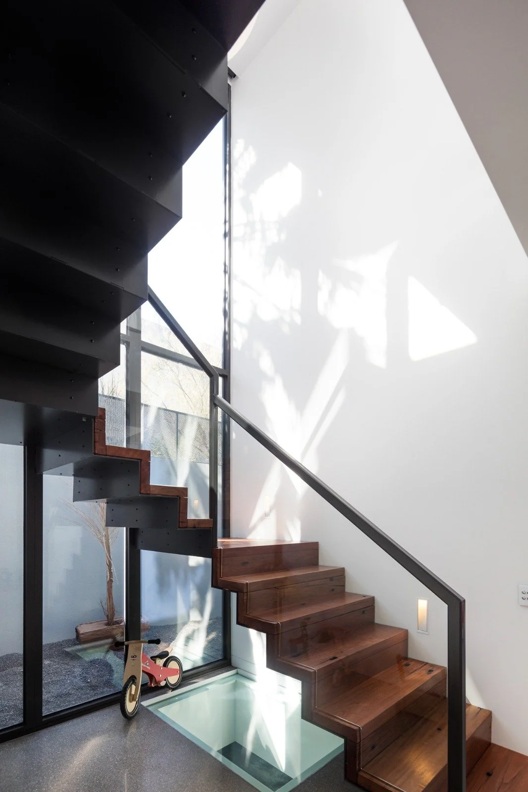 Interior view of a modern staircase with wooden steps and black metal support, next to a large glass wall letting in natural sunlight and casting shadows on the white wall.