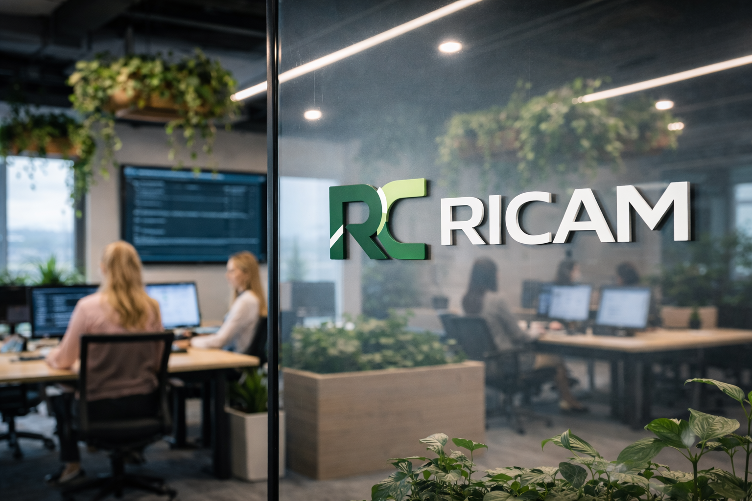 Office scene seen through a glass door with the Ricam logo, showing people working at desks with computers, greenery hanging from the ceiling, and a large screen on the wall.