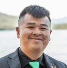 A smiling man in a dark gray suit with a turquoise tie, outdoors near a body of water with hills in the background.