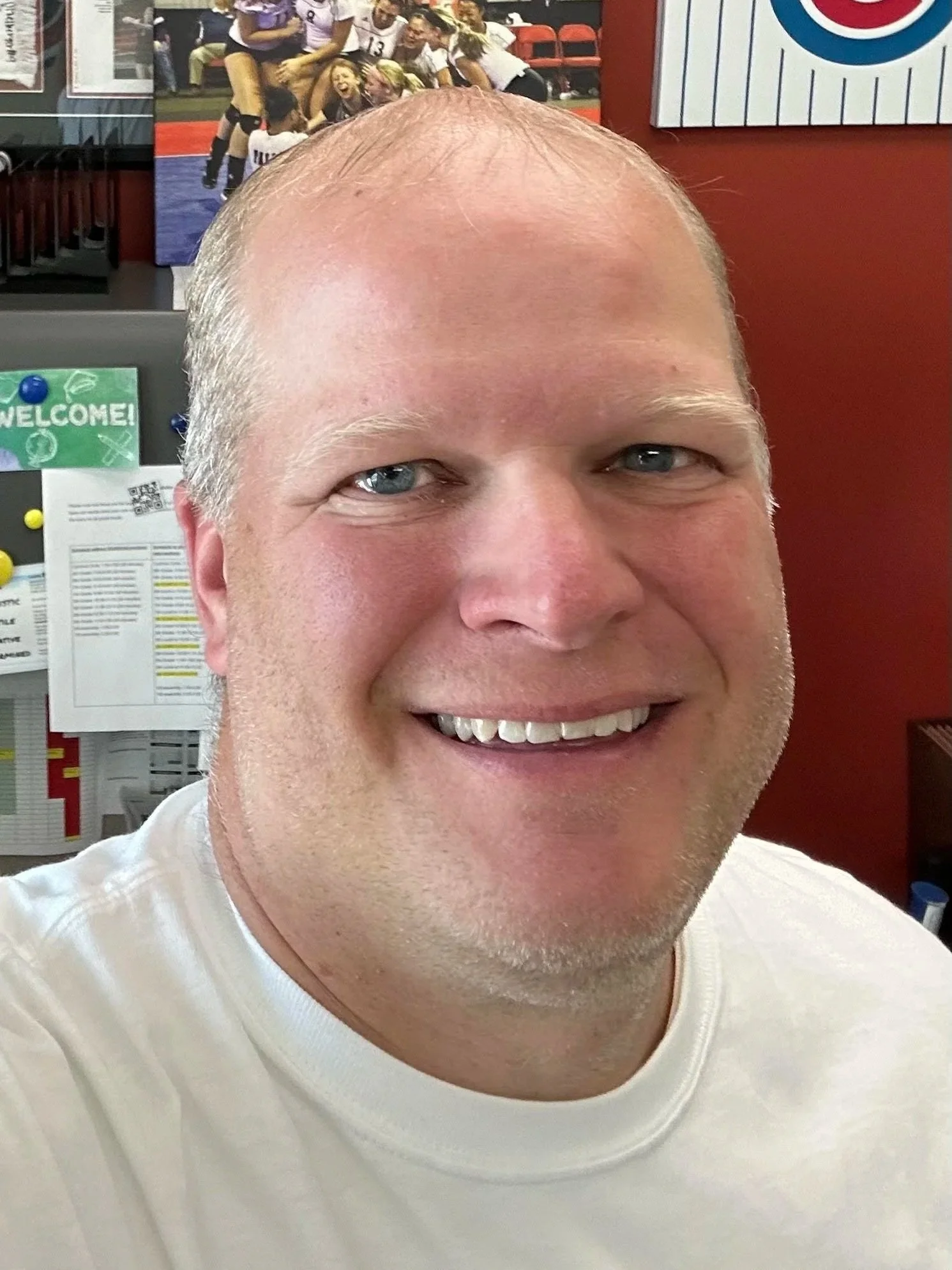 Close-up photo of smiling man with light skin and short bald head, wearing a white shirt. Background features a colorful sports-themed display with people in sports uniforms and a poster of a curling game.