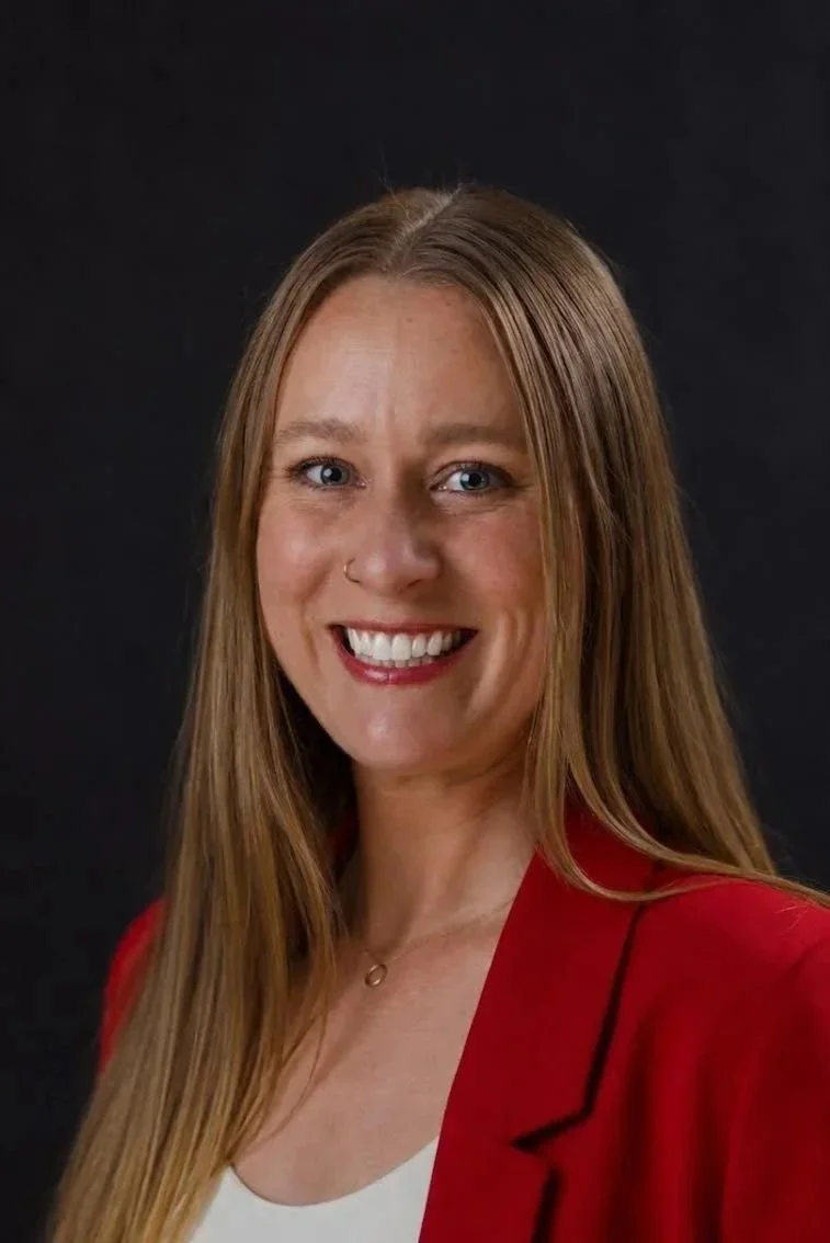 A woman with long red hair, wearing a red blazer and white top, smiling at the camera against a black background.