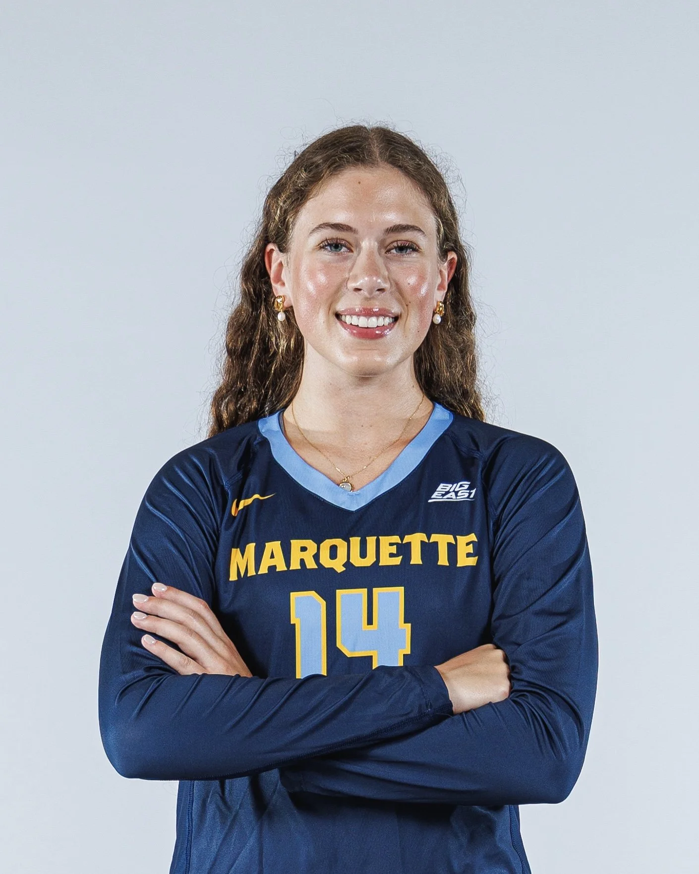 A young woman wearing a marquette jersey with the number 14, standing with arms crossed, smiling, against a light gray background.