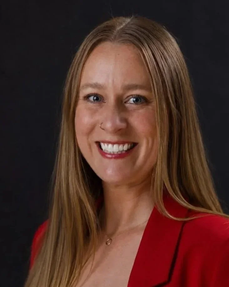 A woman with long red hair, blue eyes, and a nose piercing, smiling while wearing a red blazer against a dark background.